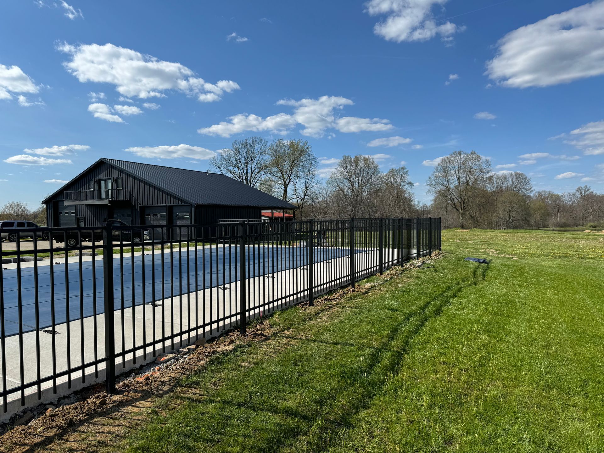 Black fence surrounds a pool and grassy area with a black building under a blue sky.