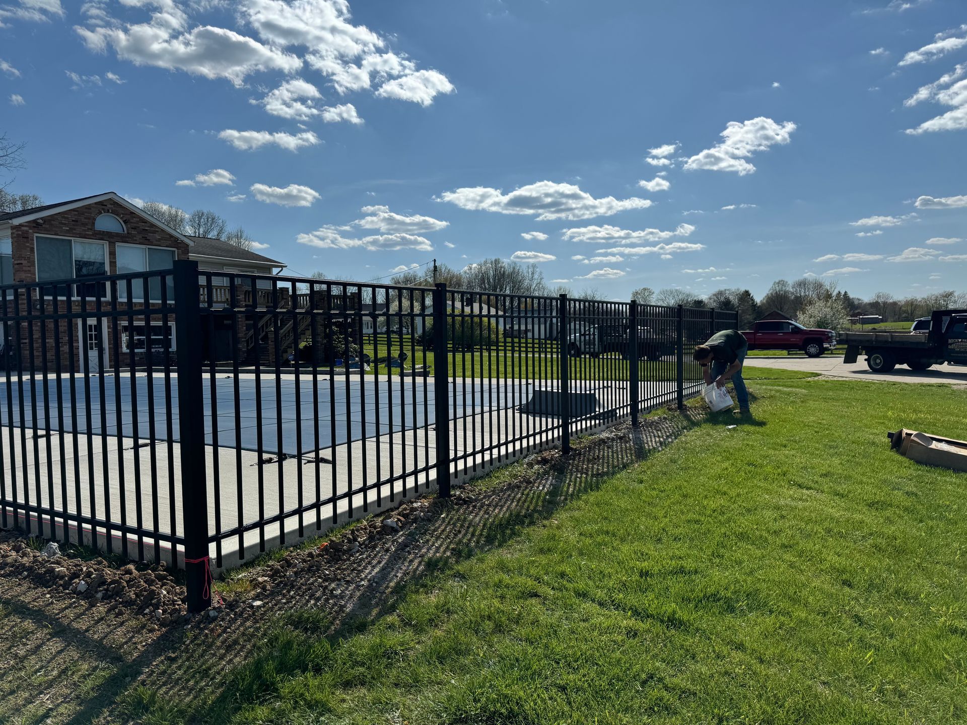 Black fence surrounds a pool, worker adjusts sections, bright sunny day.
