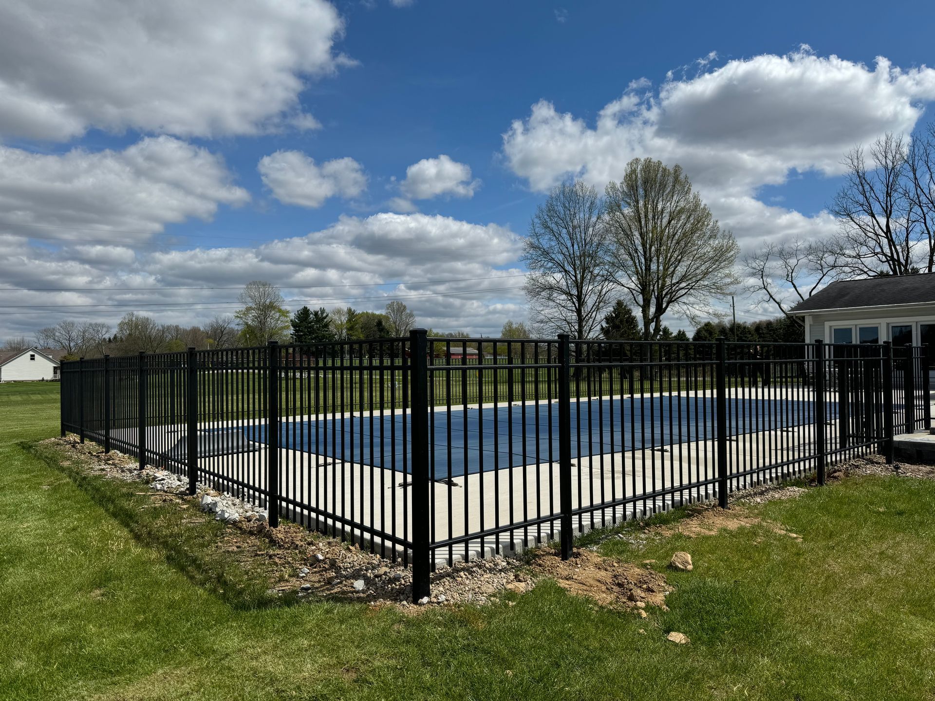 Black fenced-in pool with blue tarp, set in a grassy yard on a sunny day with white clouds.