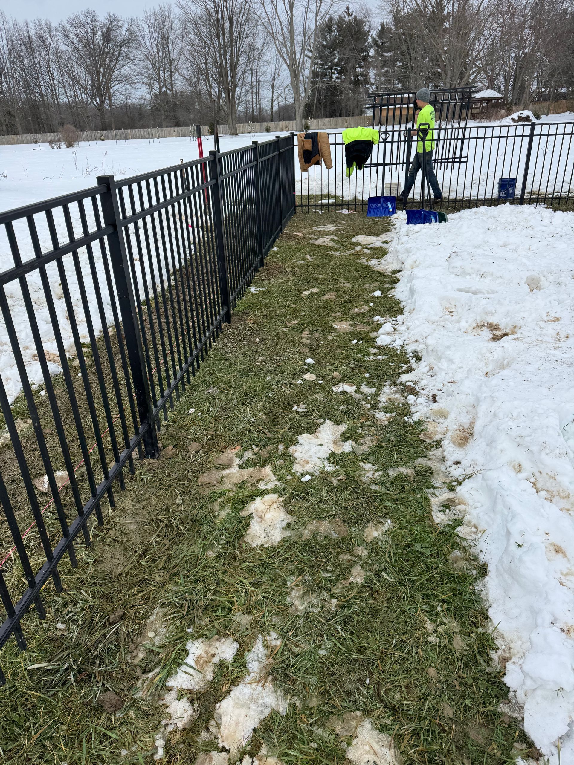 Black metal fence along grassy area with snow; person in neon vest working.
