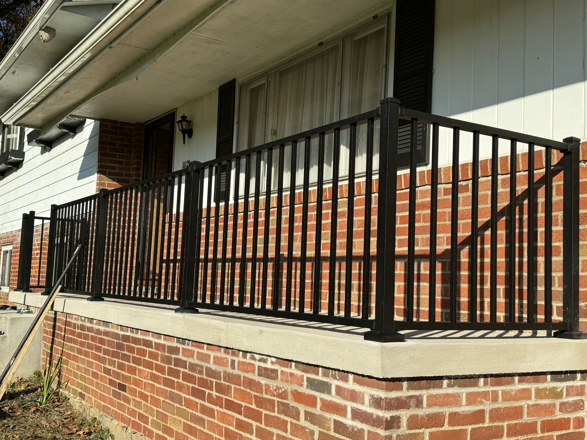 Black metal railing on a brick and concrete porch. Sunlight casts shadows.