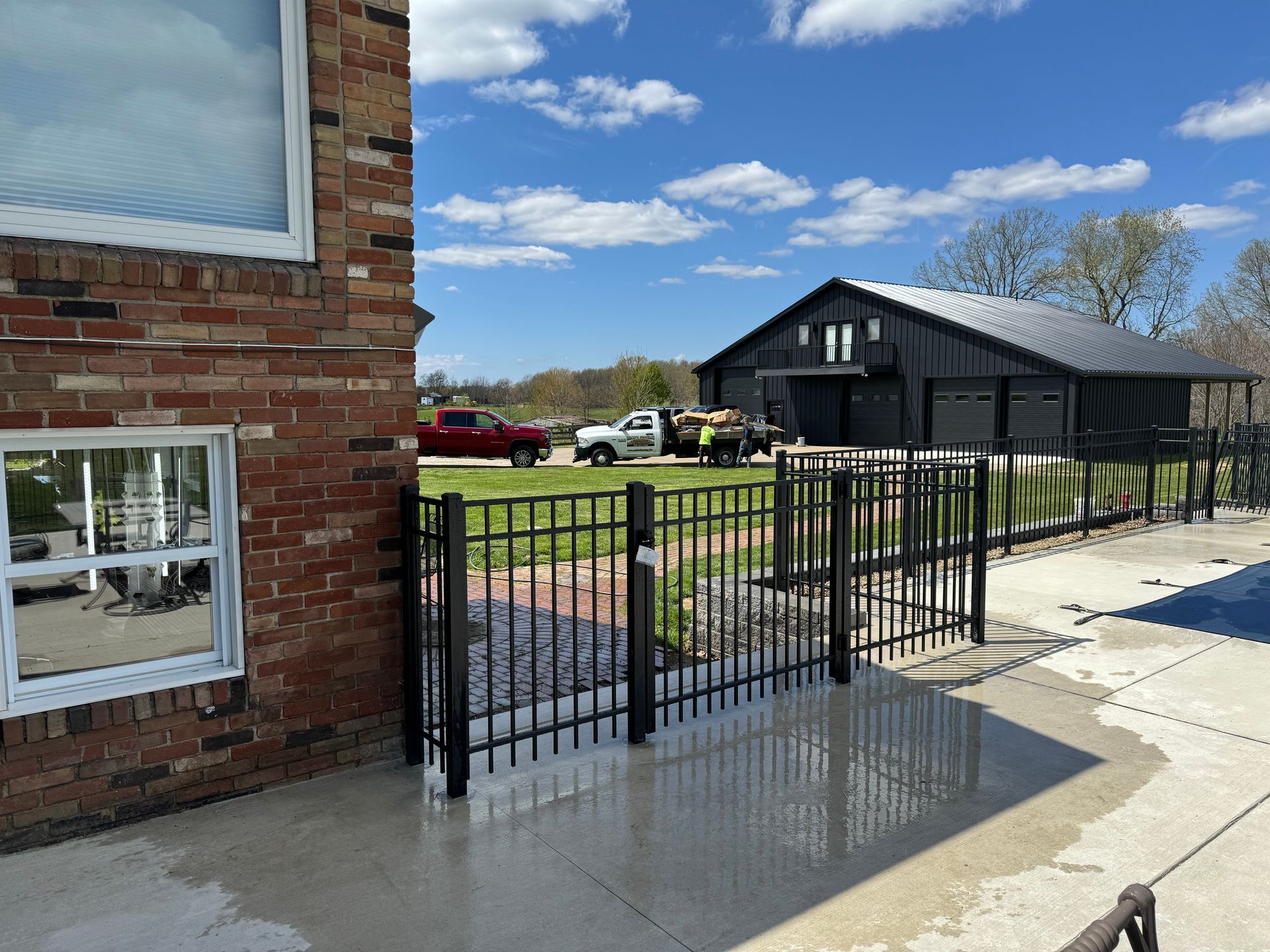 A black metal fence surrounds a pool and patio near a brick house and a black barn on a sunny day.