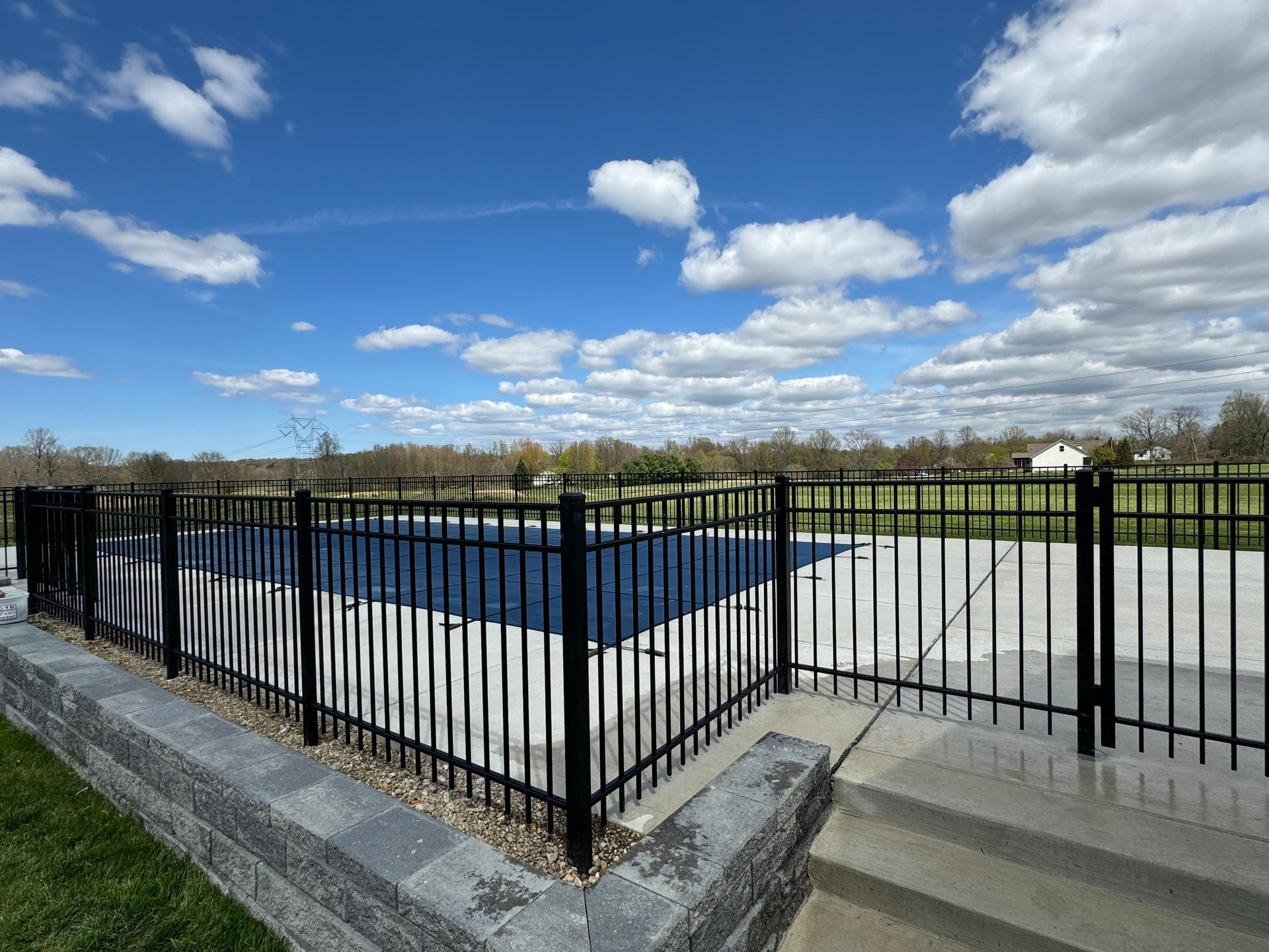 Black metal fence surrounds a pool on a sunny day. The sky is blue with scattered clouds.