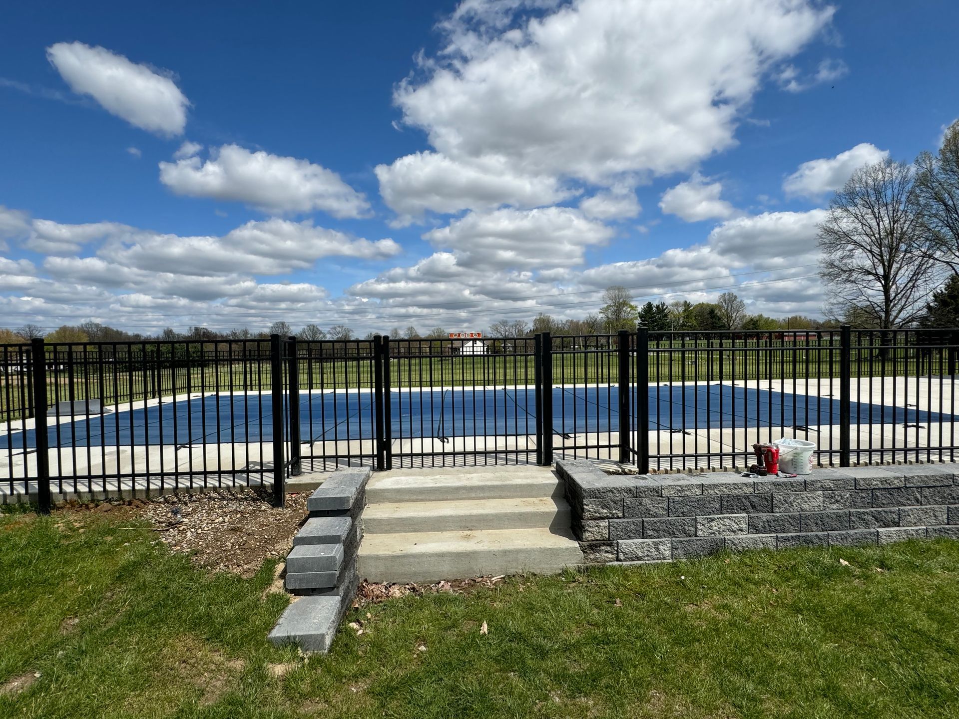 Black fenced pool with a covered surface, stone steps, and a cloudy blue sky.
