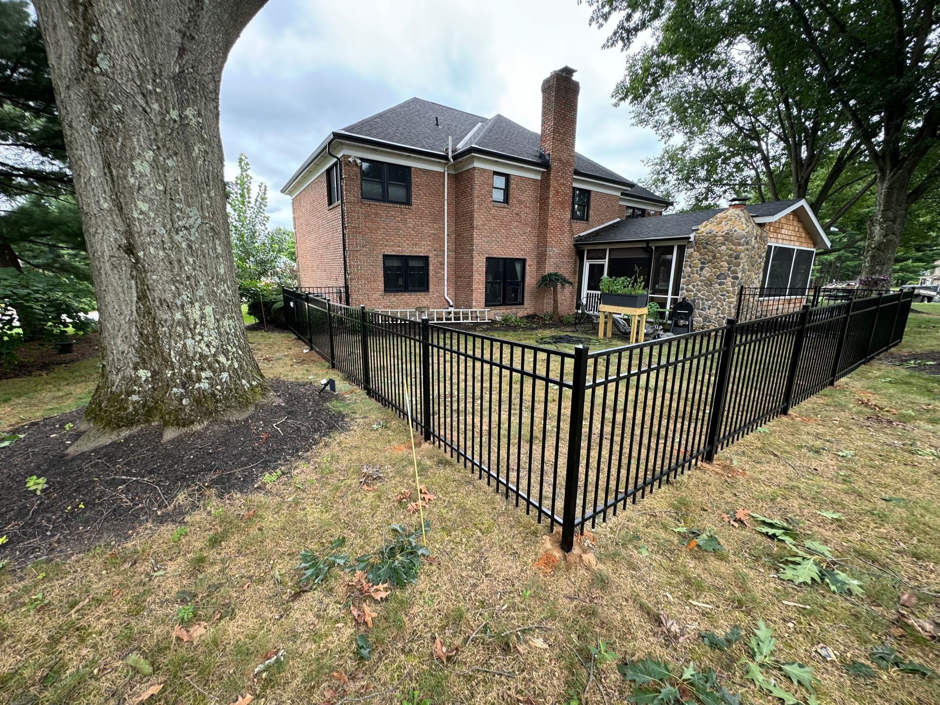 Brick house with black fence, backyard with a tree and a stone building.