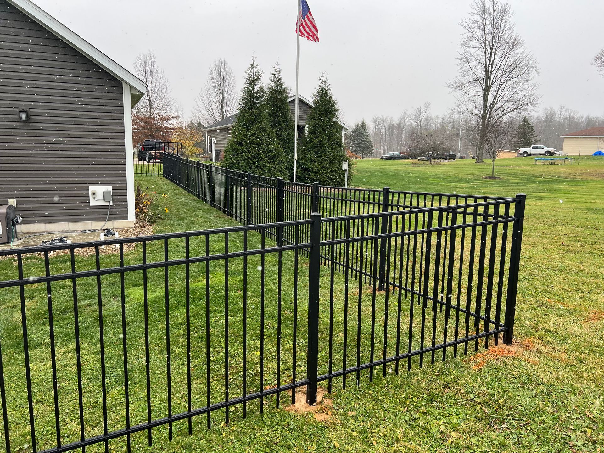Black metal fence around a yard with green grass, house on the left, American flag in background.