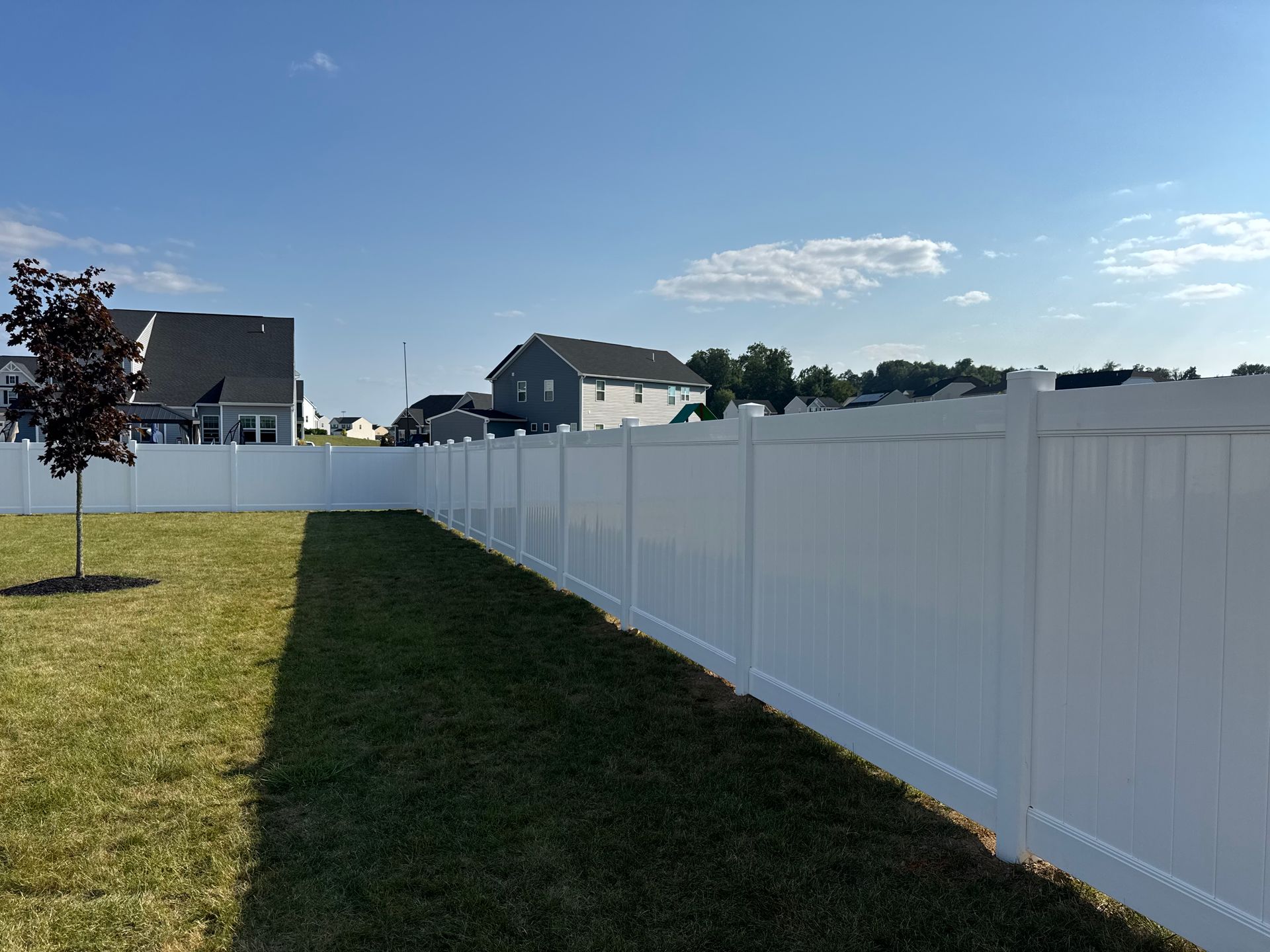 White fence bordering green lawn under a blue sky with houses in the distance.