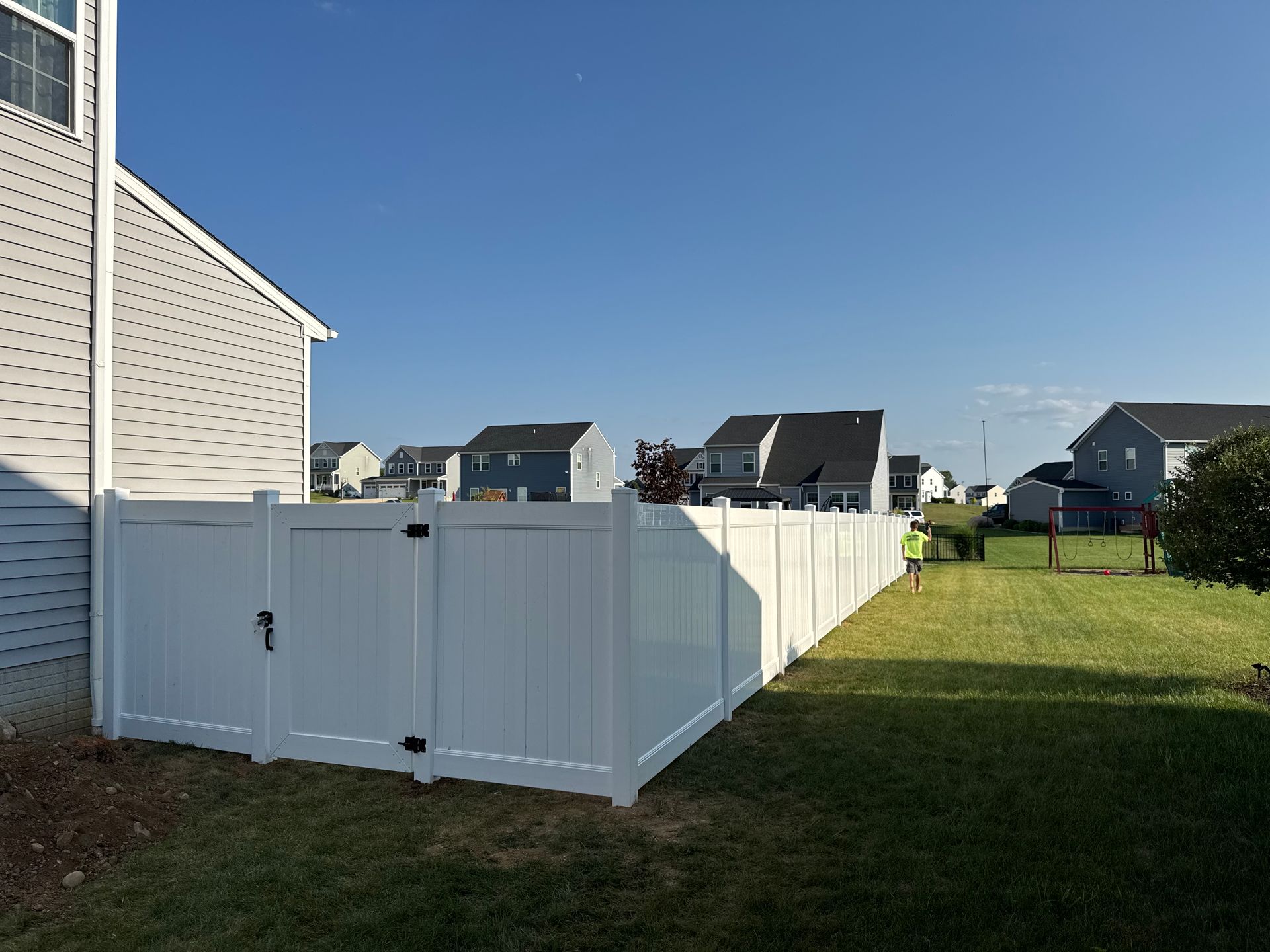 White vinyl fence surrounding a grassy yard, house on the left, sunny day.