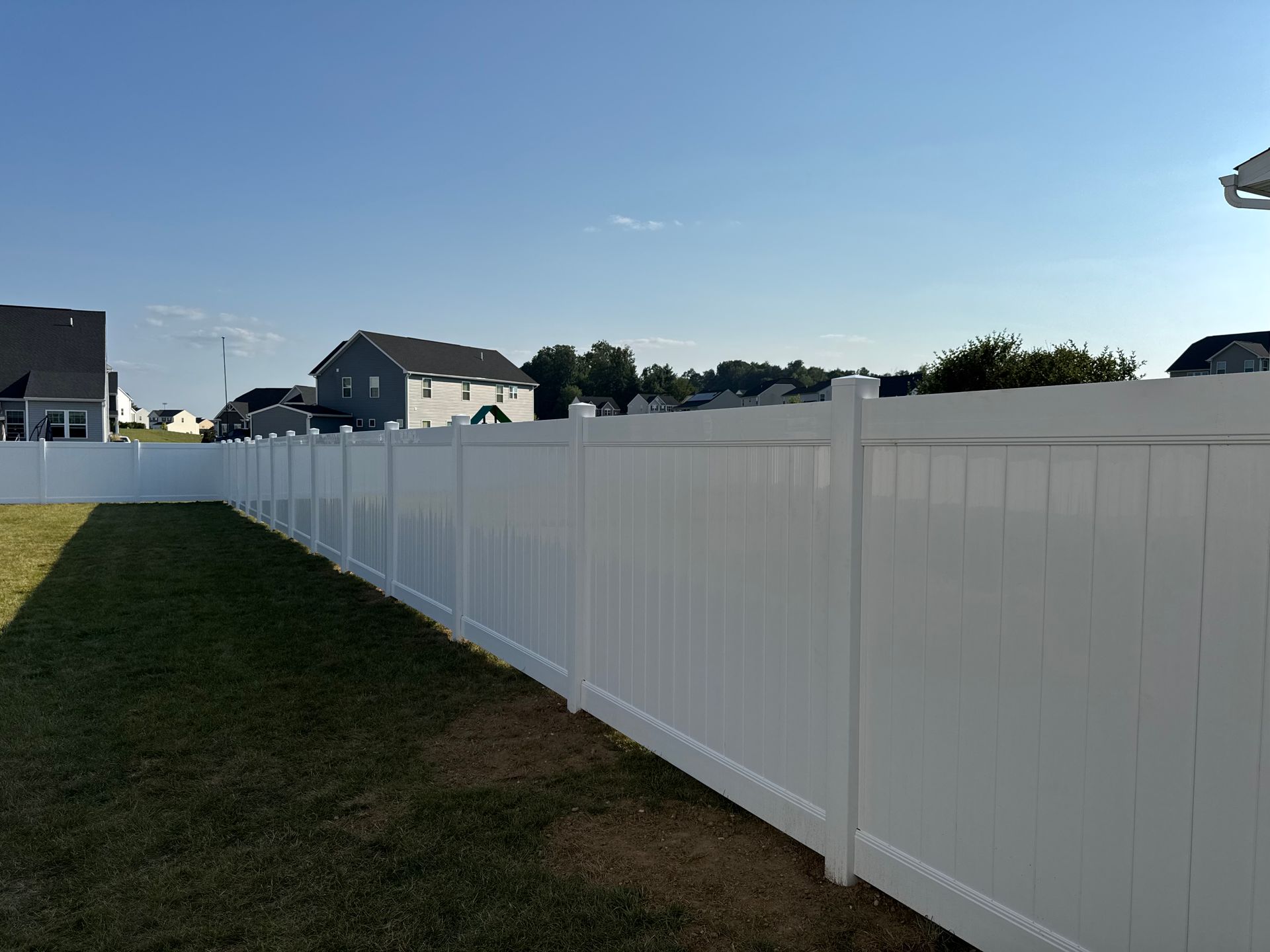 White vinyl fence in a grassy backyard, houses in the distance, blue sky overhead.