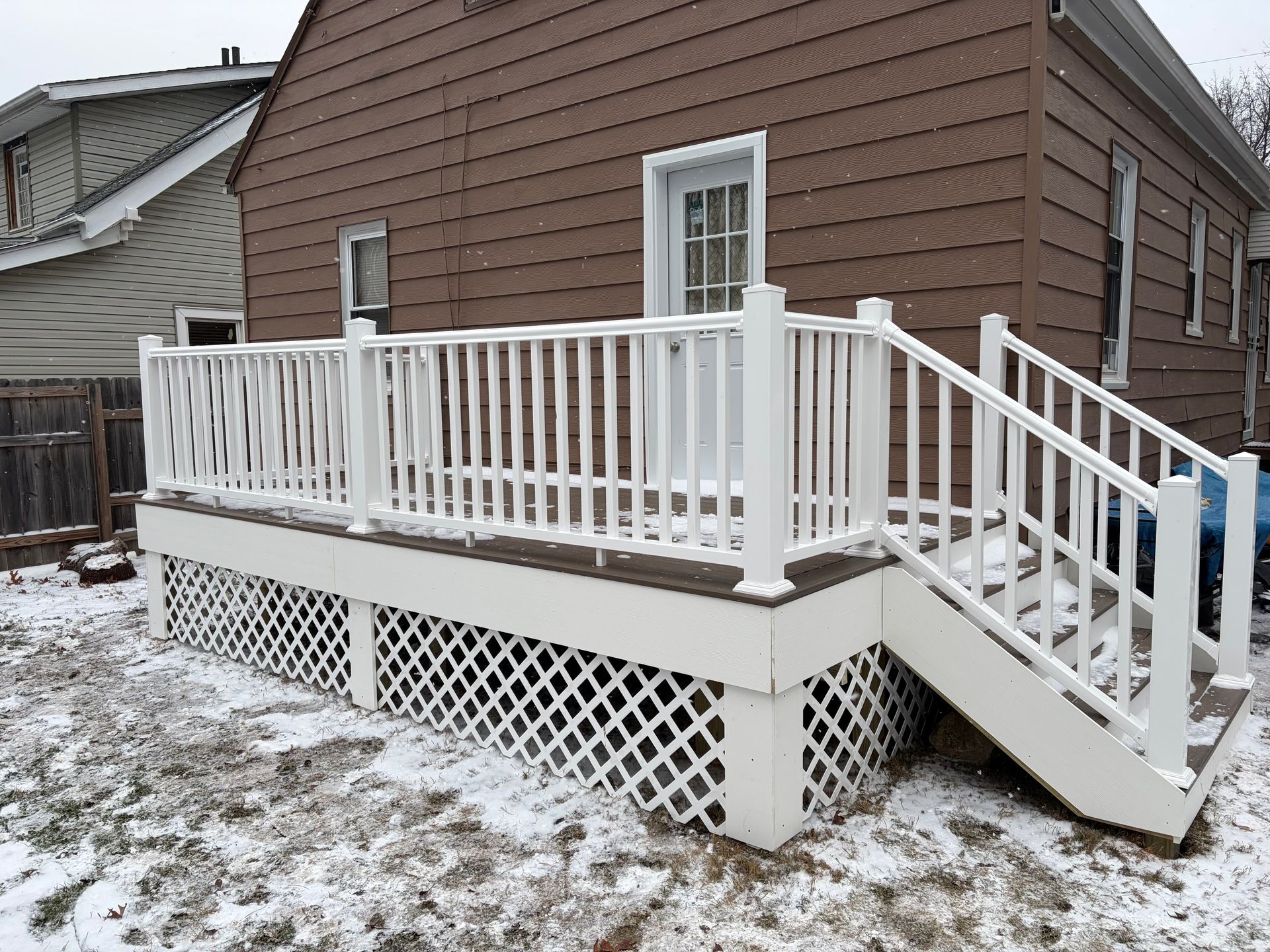 White deck with lattice skirting and railing, stairs, and brown siding on a house, snow on the ground