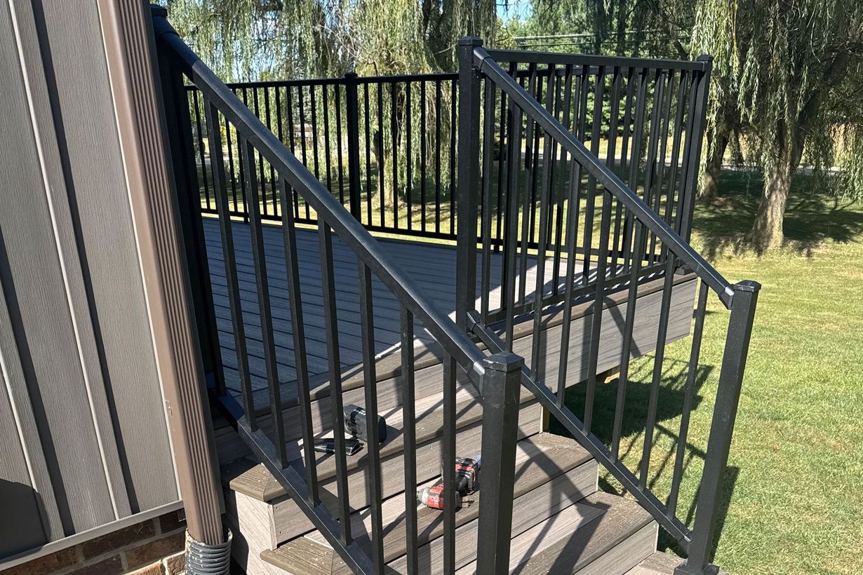 Black deck with stairs, railing, and a green lawn in the background.