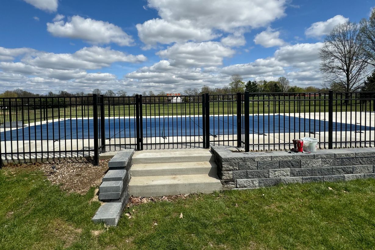 Black fence surrounds a blue-covered pool. Steps lead to a gate in the fence. Overcast sky.