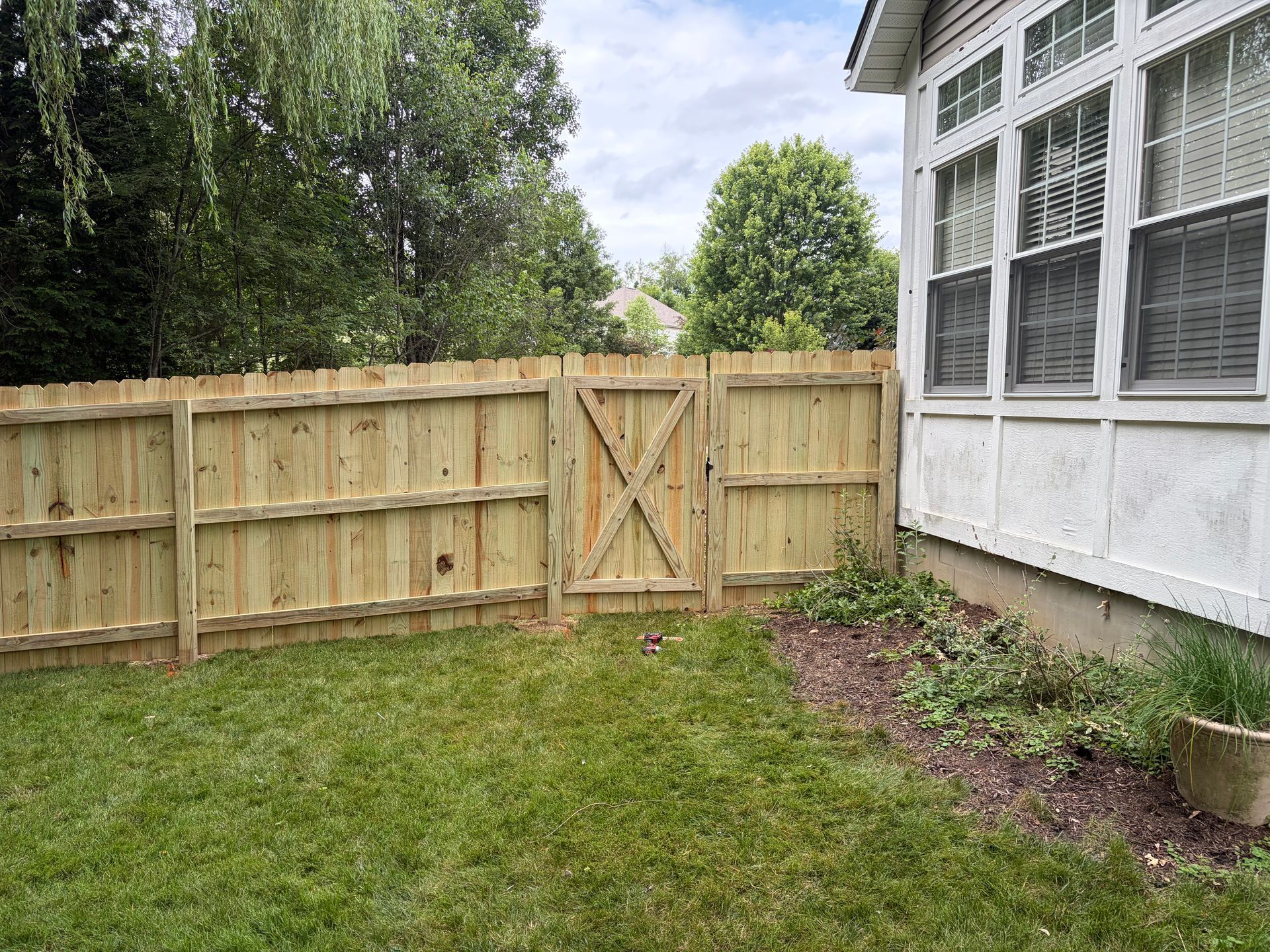 Backyard with a wooden fence gate, green lawn, and a white house with windows on the right.