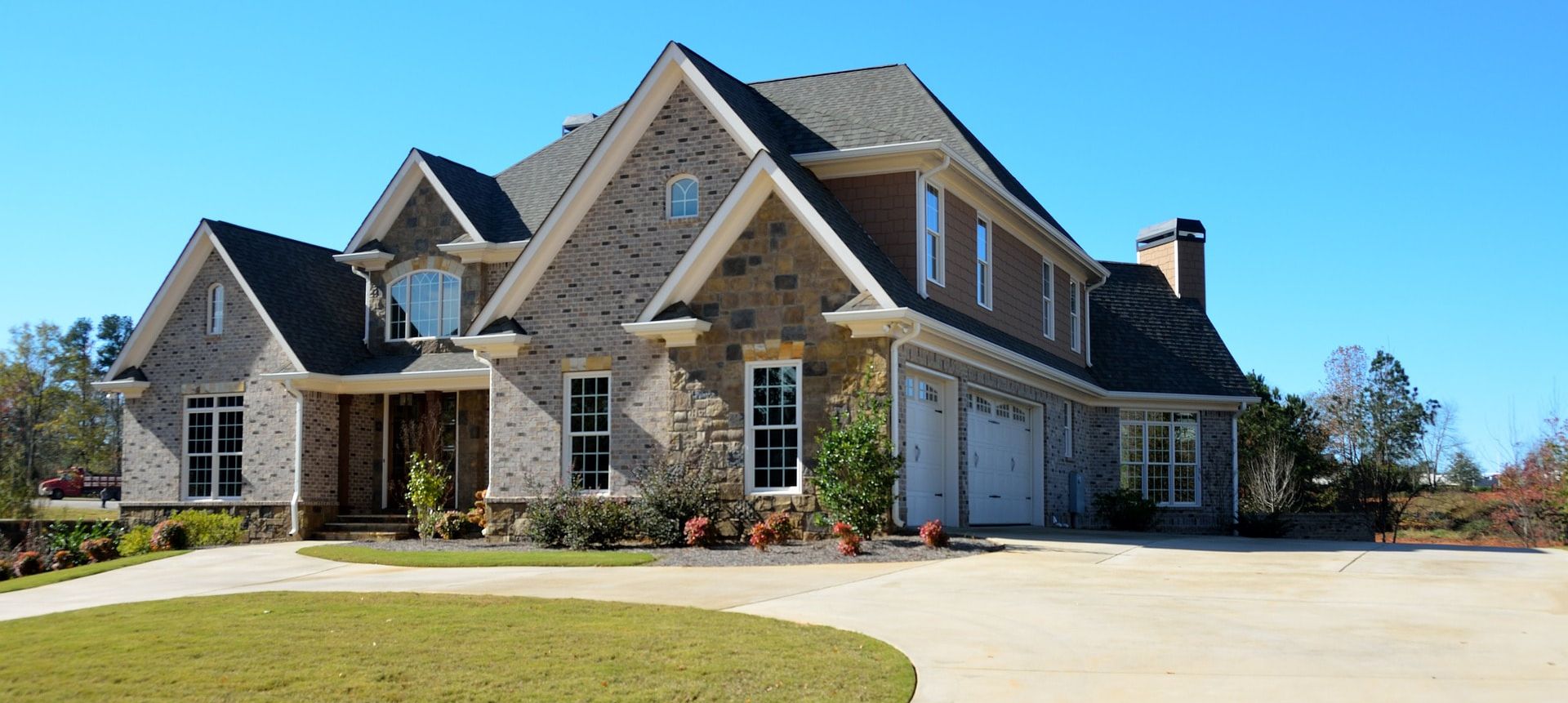 Stone suburban house with a two-car garage, steep roof, and a curved driveway under a clear blue sky