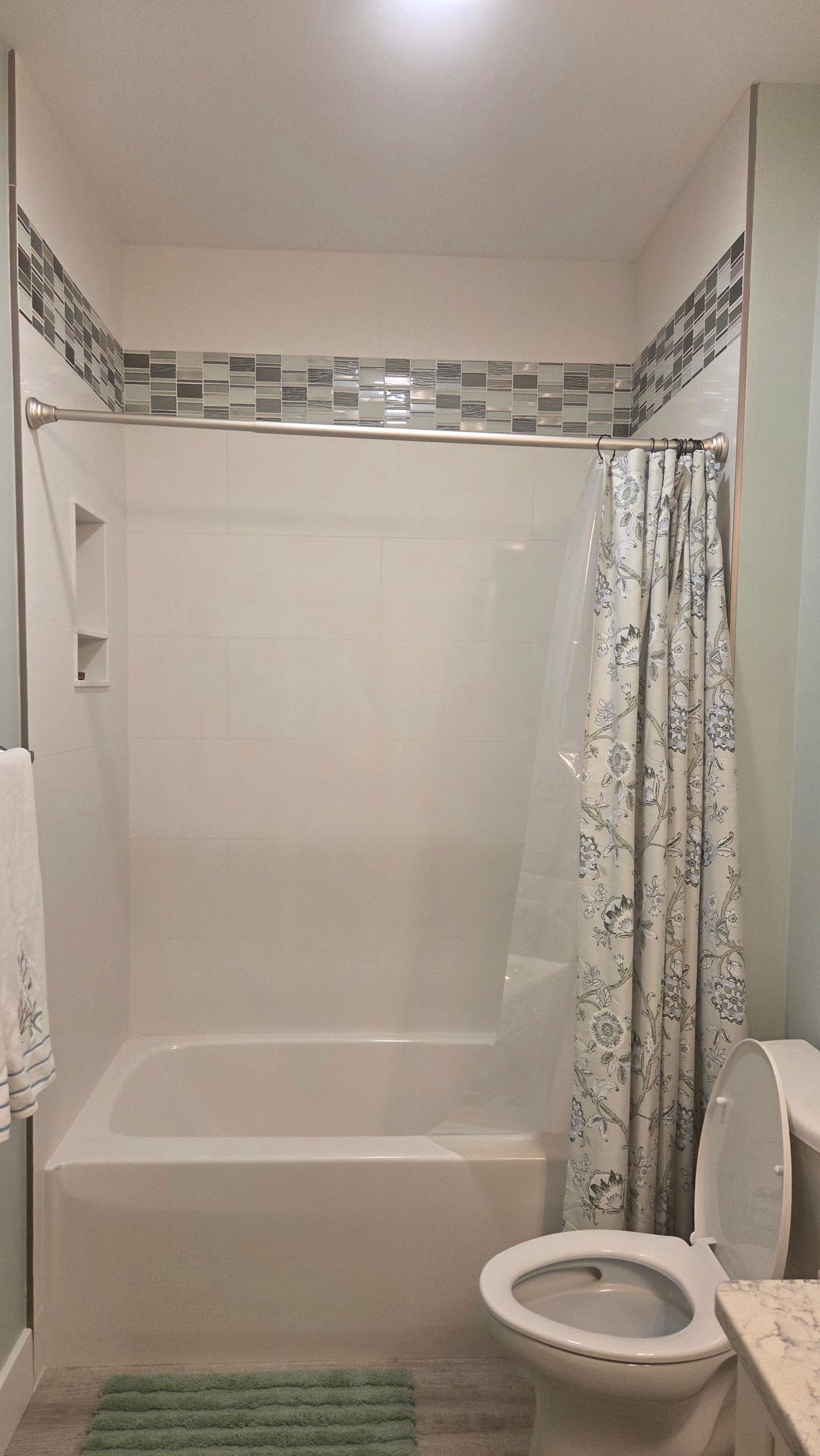 Small bathroom with white tub-shower, mosaic border, patterned curtain, and toilet beside vanity.