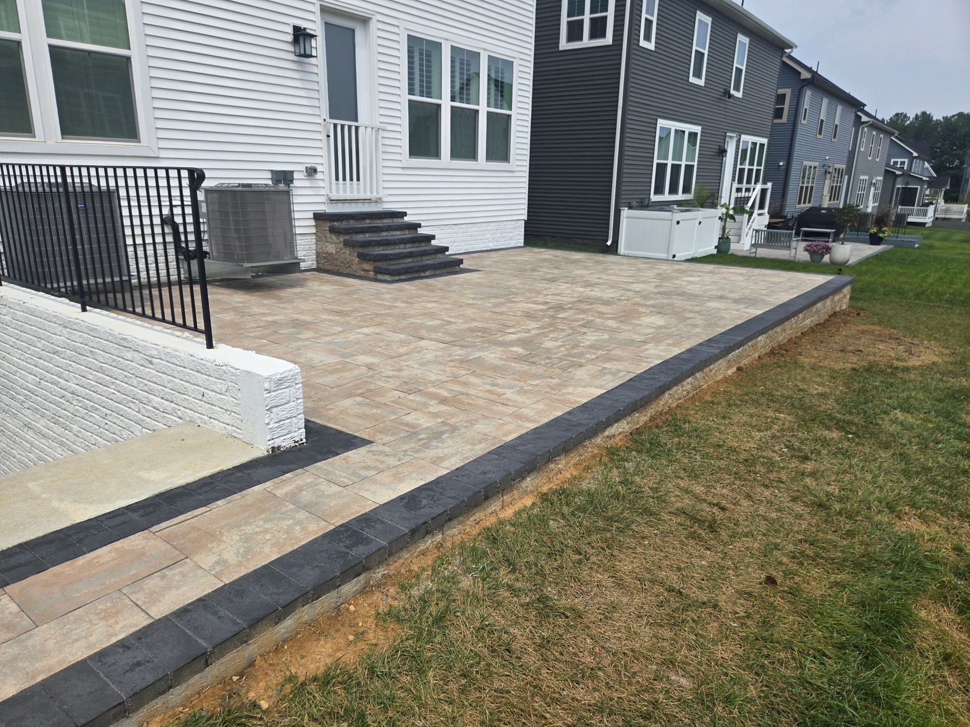 Paved patio beside row houses, with black railings and a grassy lawn edge.