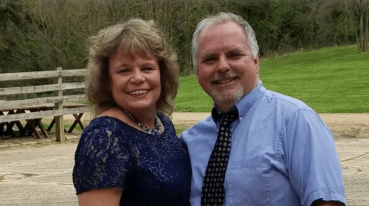 A smiling couple standing side-by-side outdoors, the woman in a navy lace top and the man in a blue shirt and patterned tie.