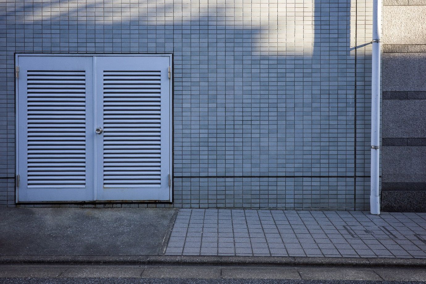 White louvered doors on a blue-tiled wall, beside a gray tiled sidewalk and a metal pole.