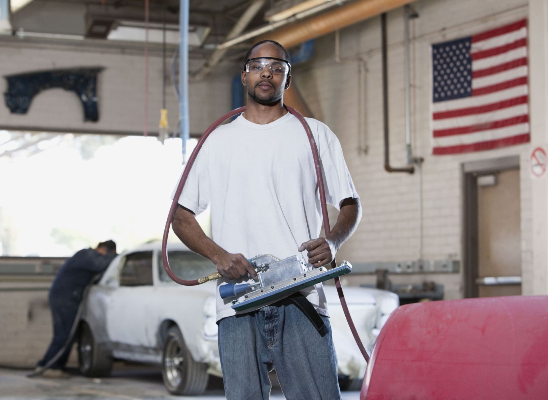 Man holding a circular saw in a garage workshop beside a car, with an American flag on the wall.