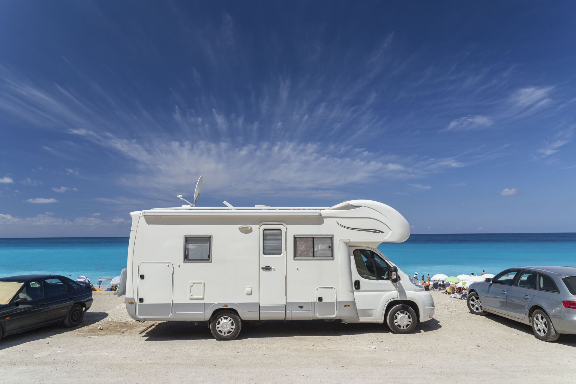 White RV parked on a beach, blue water and sky in the background. Two cars are parked on either side.