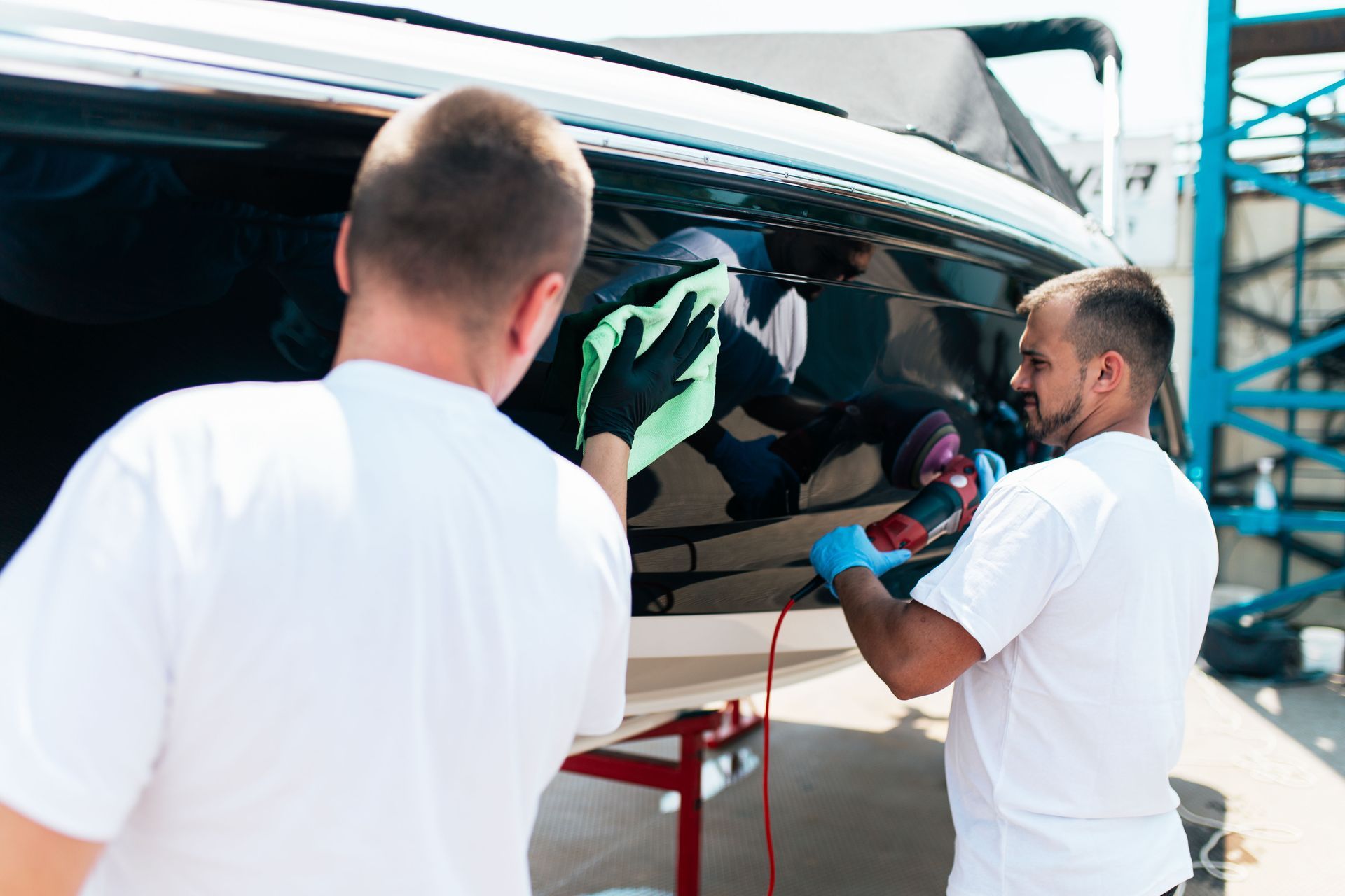 Two people cleaning a black boat hull: one wiping with a green cloth, the other using a buffer.