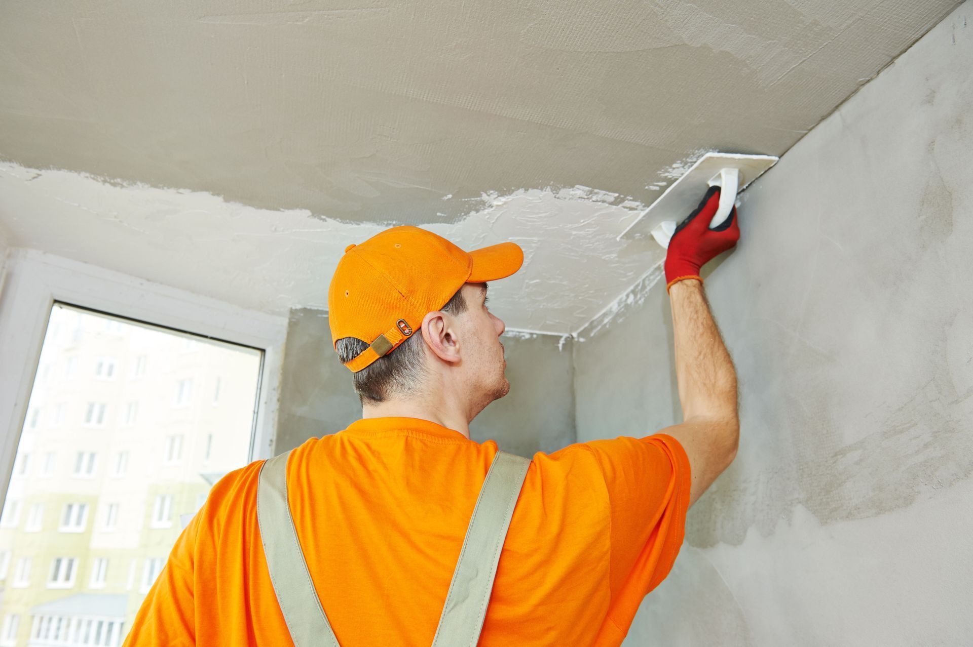 Man in orange shirt and cap plastering a ceiling with a trowel, indoors.