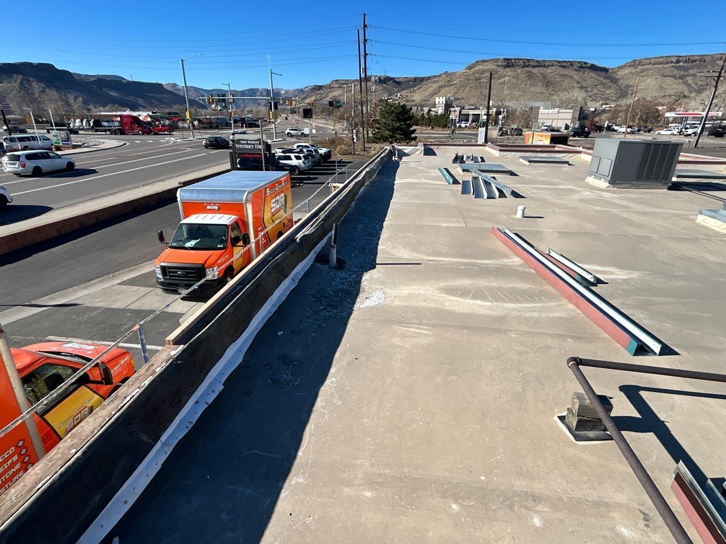 An orange moving truck on a road next to a building's rooftop, sunny day. Mountains in background.