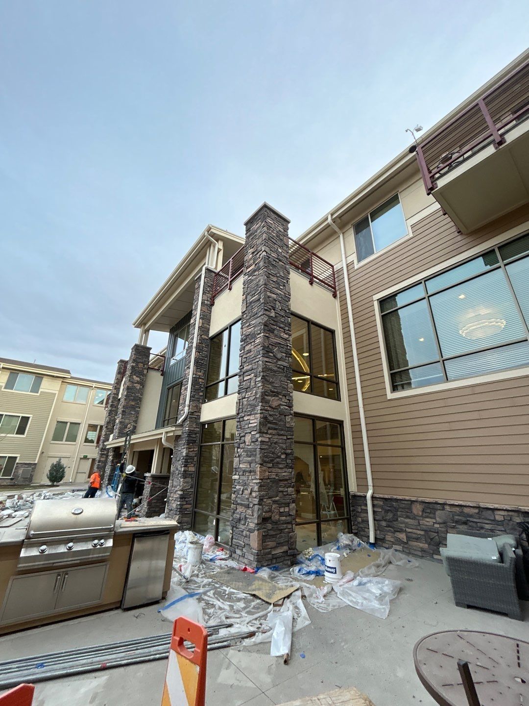 Outdoor view of a multi-story building facade with stone pillars, large windows, and patio furniture under construction.