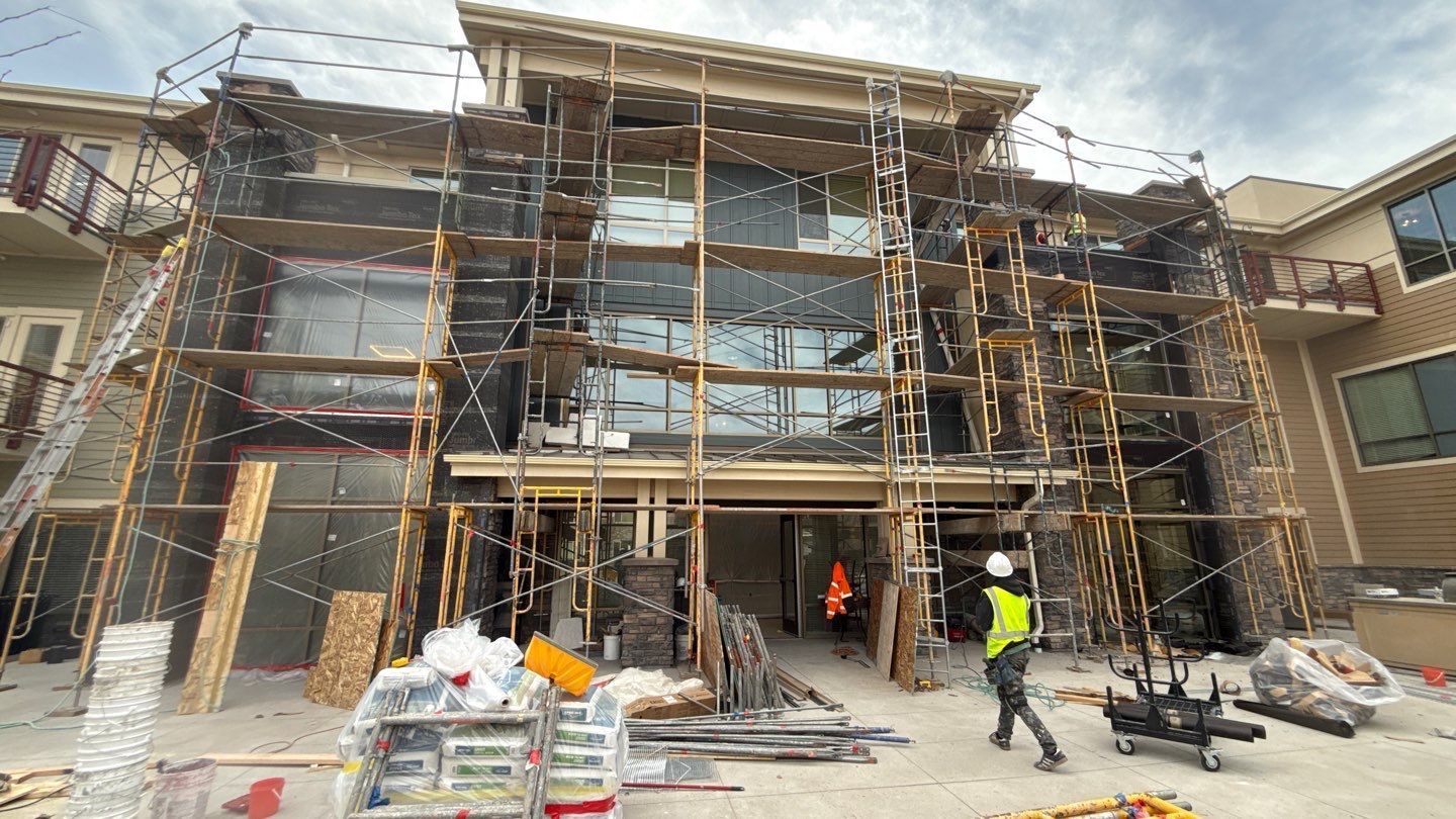 A construction worker in a high-visibility vest stands in front of a multi-story building covered in wooden scaffolding.