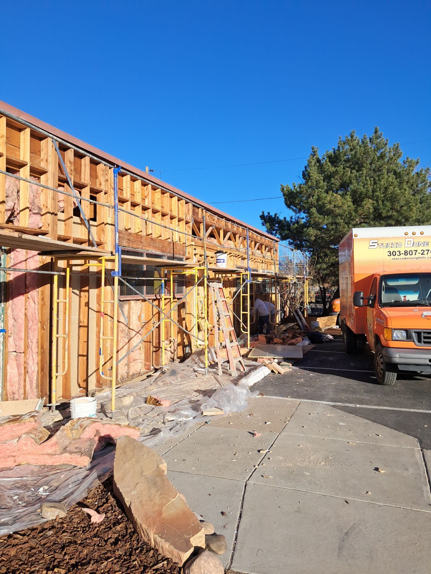 Building under construction; wood framing, scaffolding, and orange moving truck on a sunny day.
