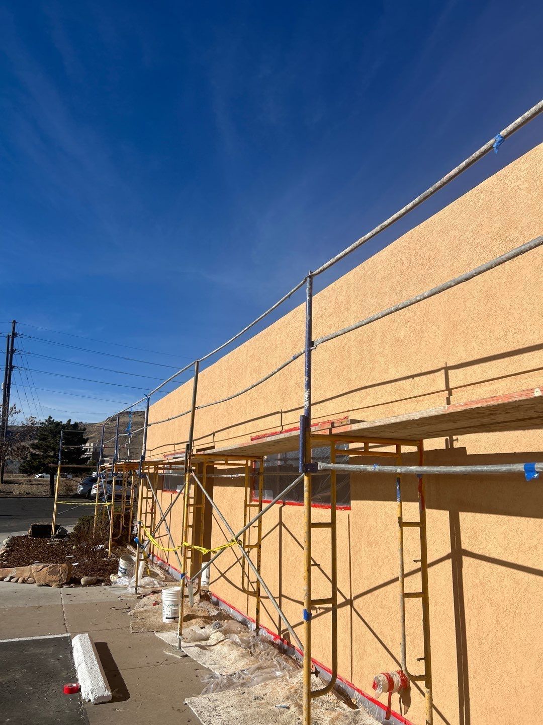Scaffolding on building exterior under clear blue sky during construction. Yellow stucco, tan boards.