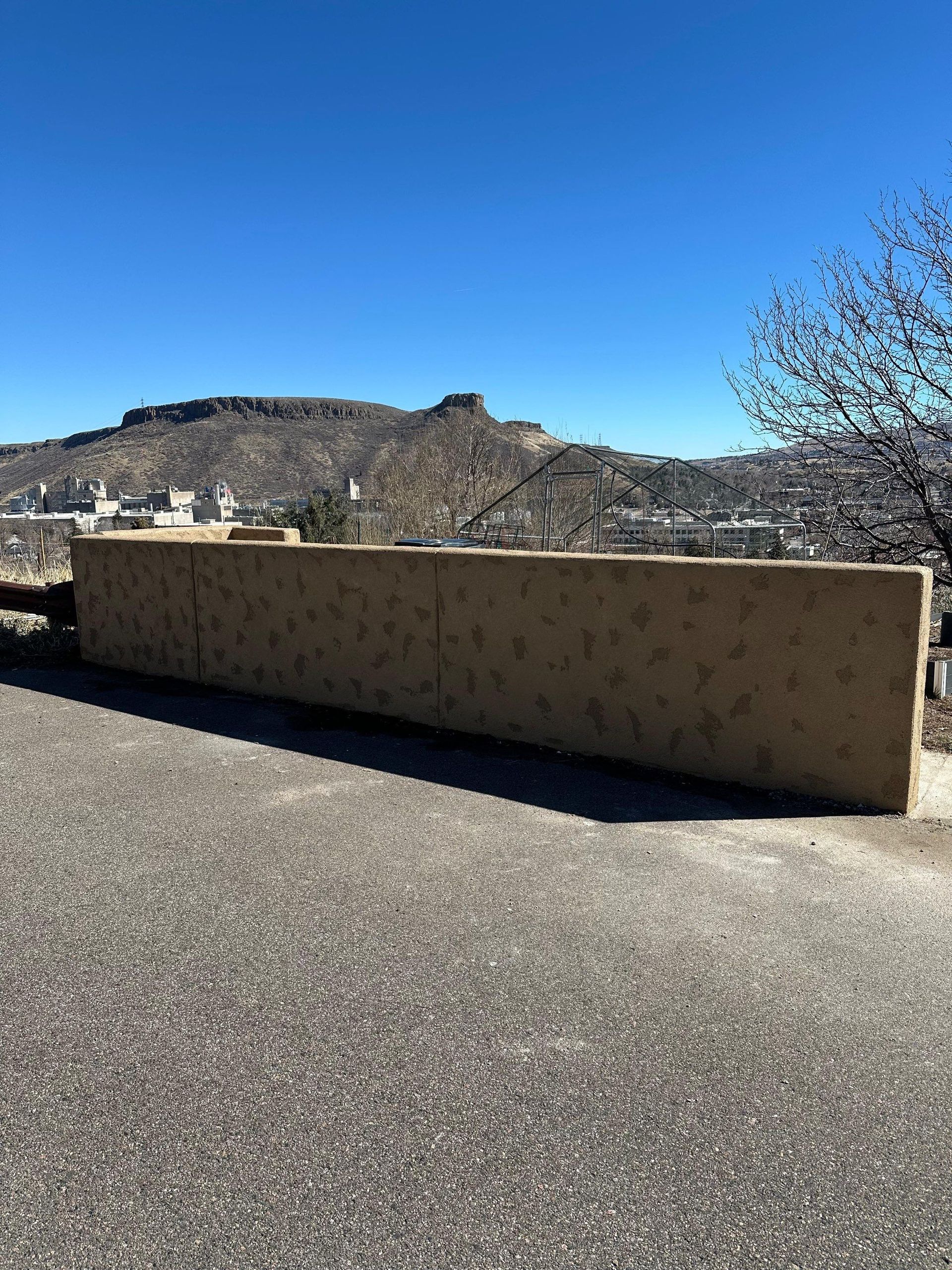 A tan textured retaining wall sits beside an asphalt road, overlooking a valley with a mountain under a clear blue sky.