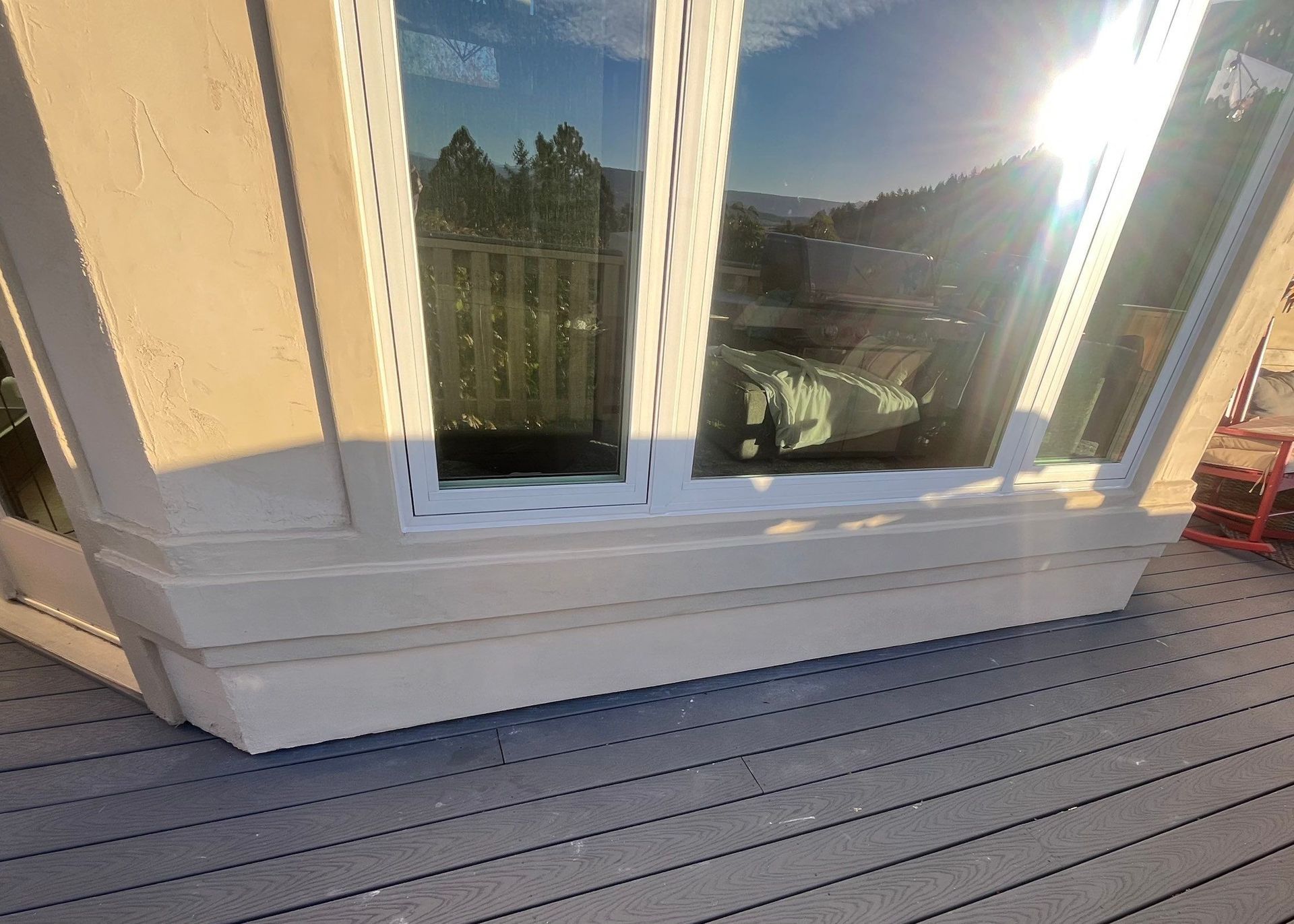 Exterior view of a window with white trim, reflecting a landscape, on a beige-colored structure, with gray decking. 