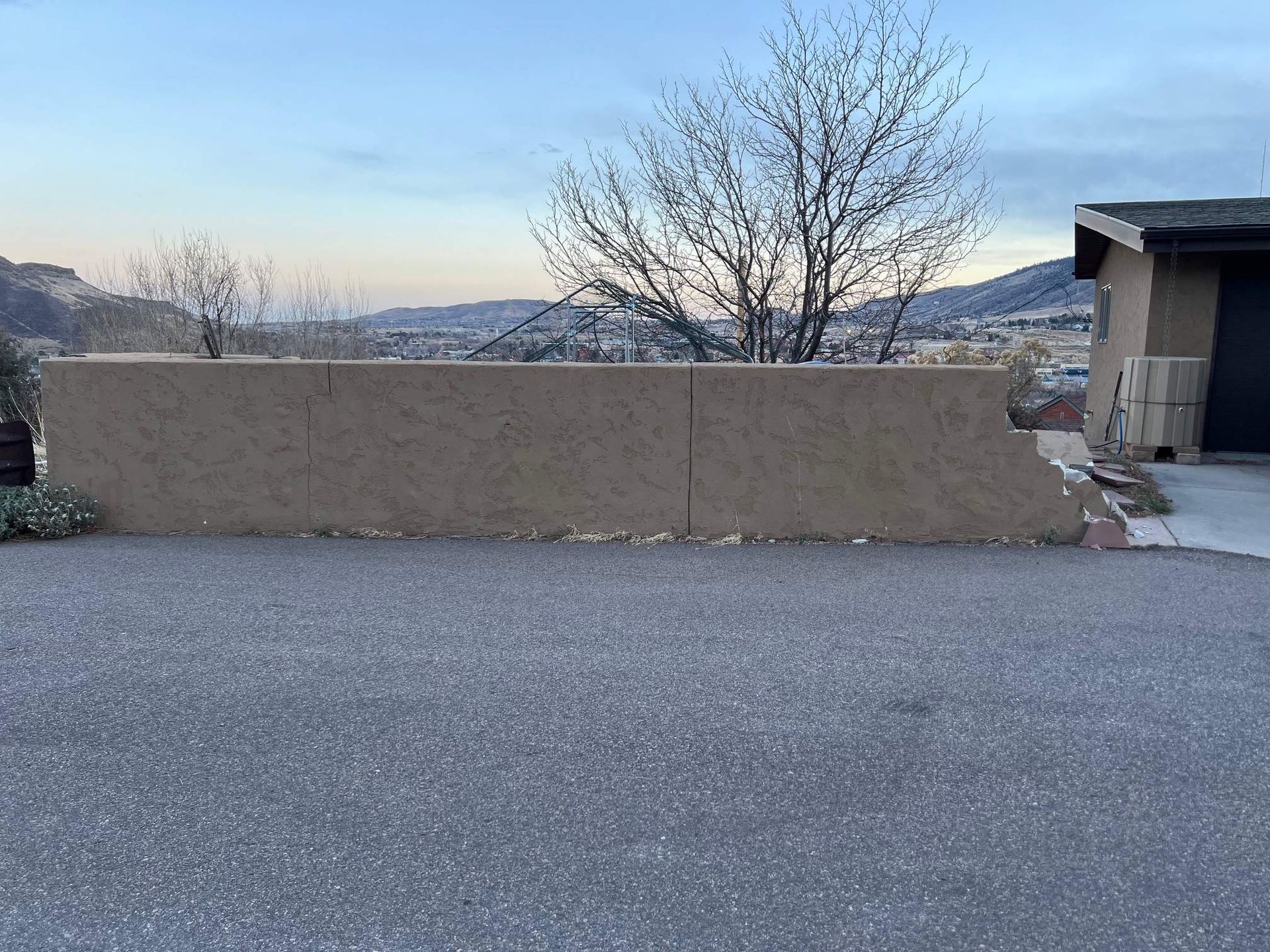 A tan adobe-style wall borders a gravel driveway in front of a house, with distant hills visible under a blue sky.