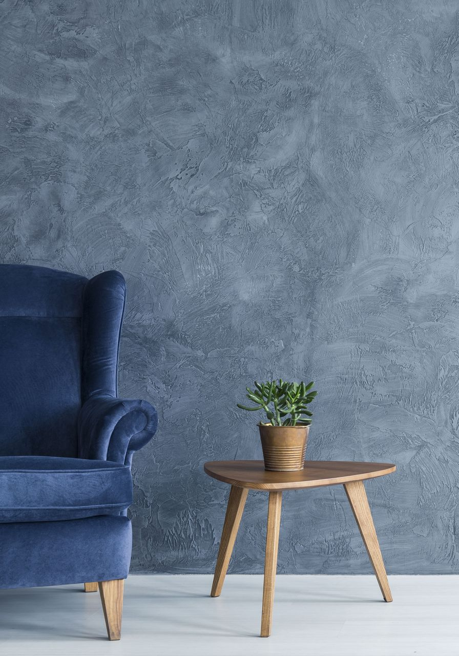Blue armchair, small wooden table with potted plant, against textured gray wall, white floor.