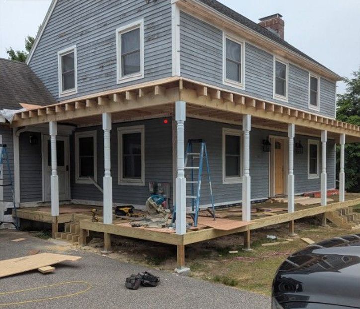A two-story house with a wrap-around porch under construction; a worker uses a saw.