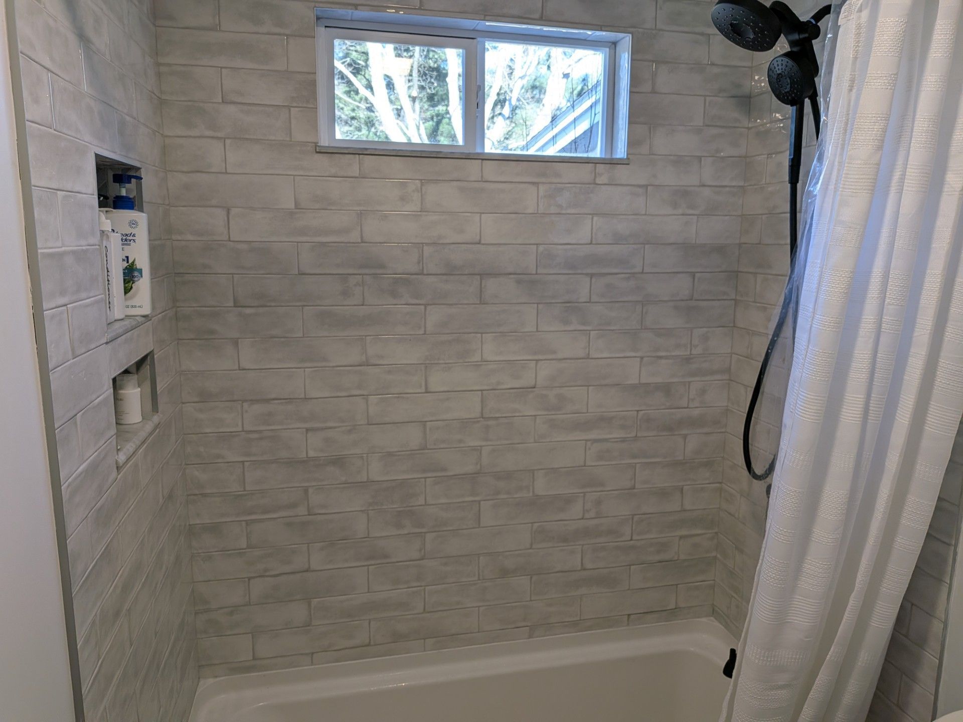 Shower with gray brick-like tile, small window, black showerhead, and a white shower curtain.