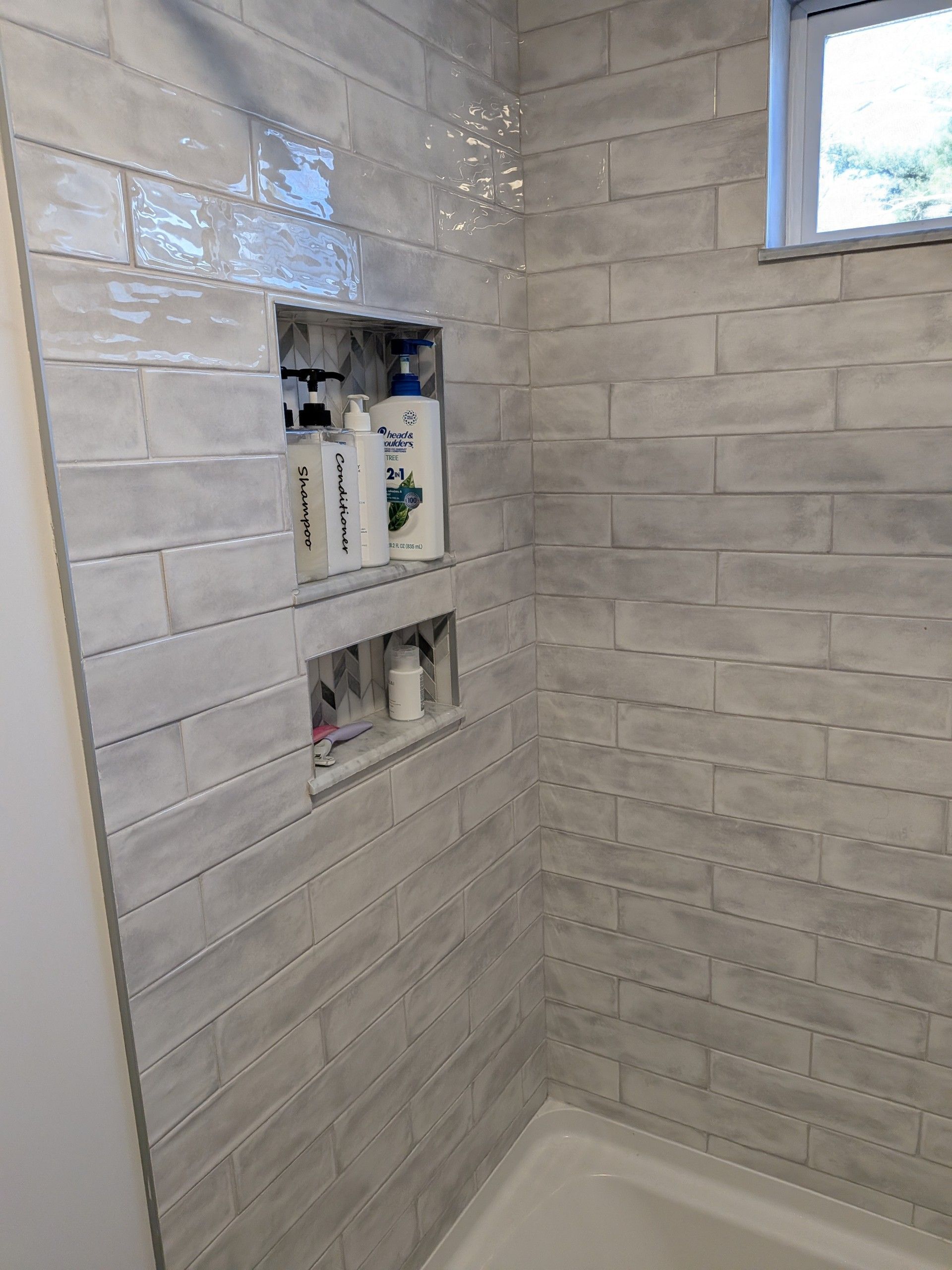 Shower with gray brick-like tiles, two built-in shelves with toiletries, and a small window.