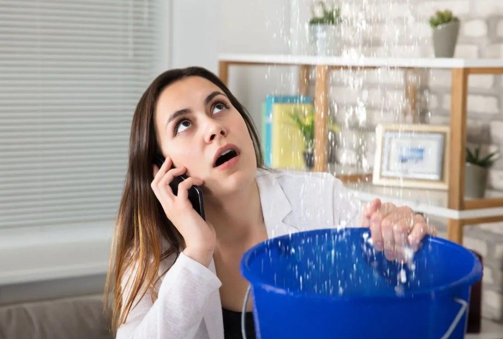 Woman on phone, catching water leaking from ceiling in blue bucket. Interior setting.