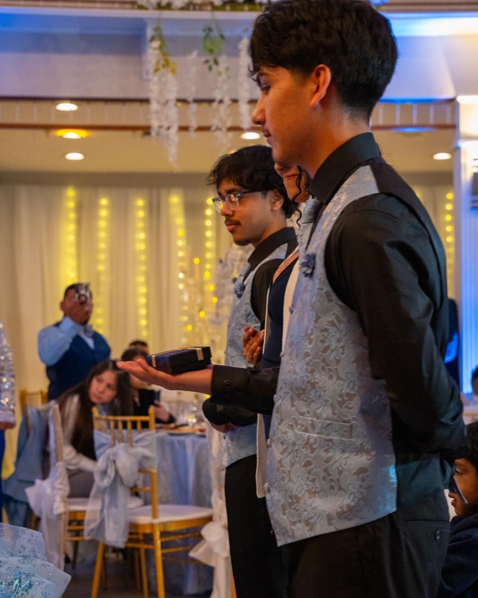 Three young men in vests stand at a formal event, one gestures with an open palm.