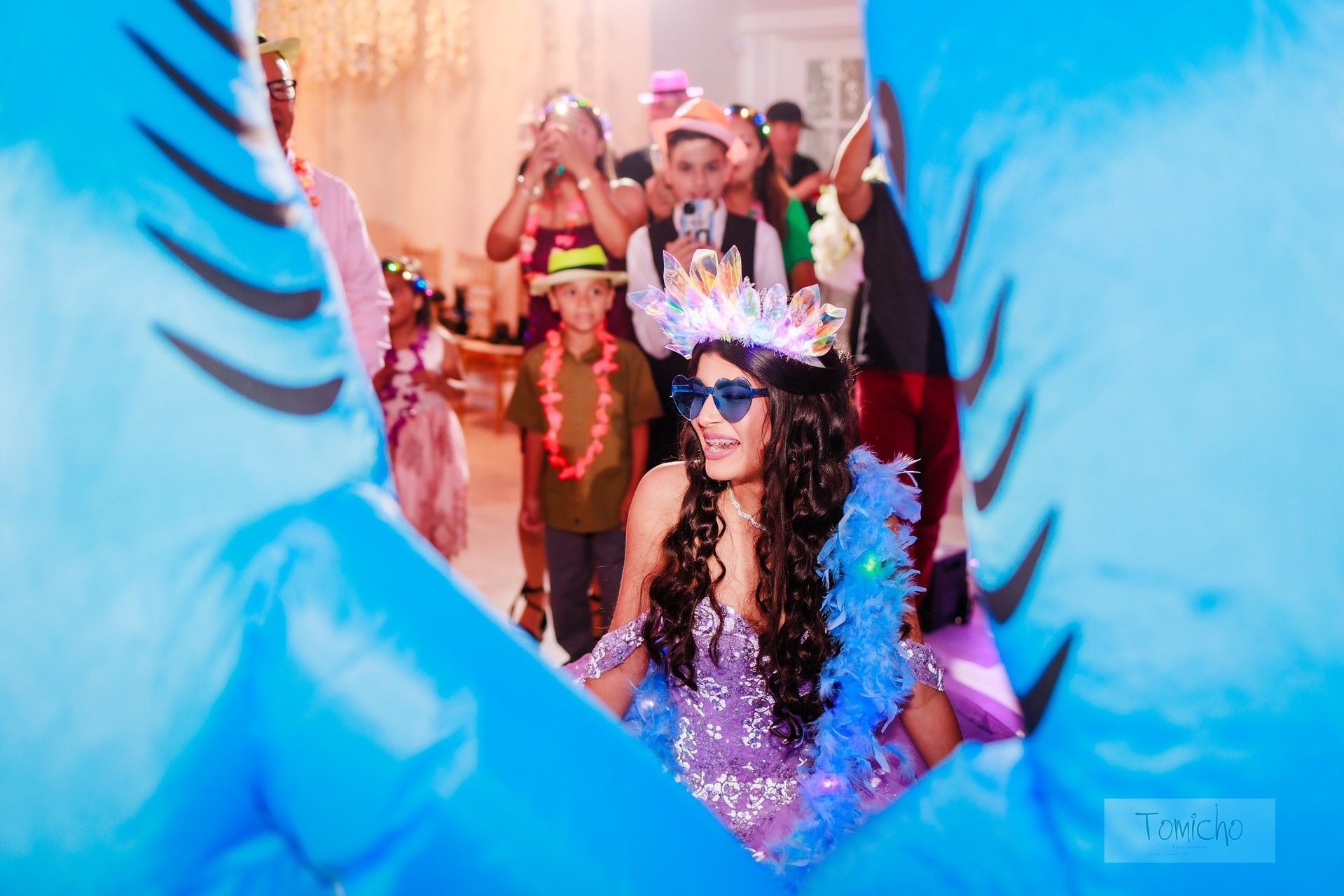 Teen in a sparkly dress and crown, surrounded by partygoers, visible through a blue cutout.