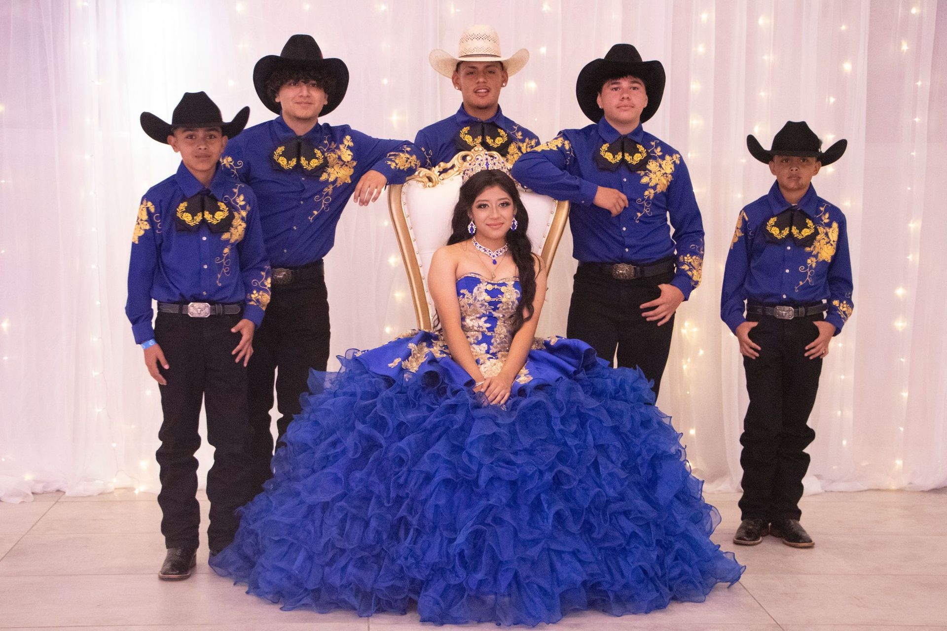 A young person in a blue formal gown sits with five people in blue shirts, black hats, and black pants.