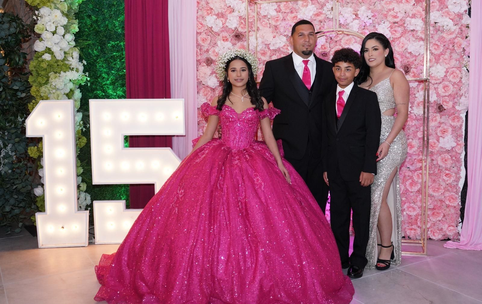 Family posing at a quinceañera. The girl in a pink dress, parents, and brother. Pink floral backdrop.