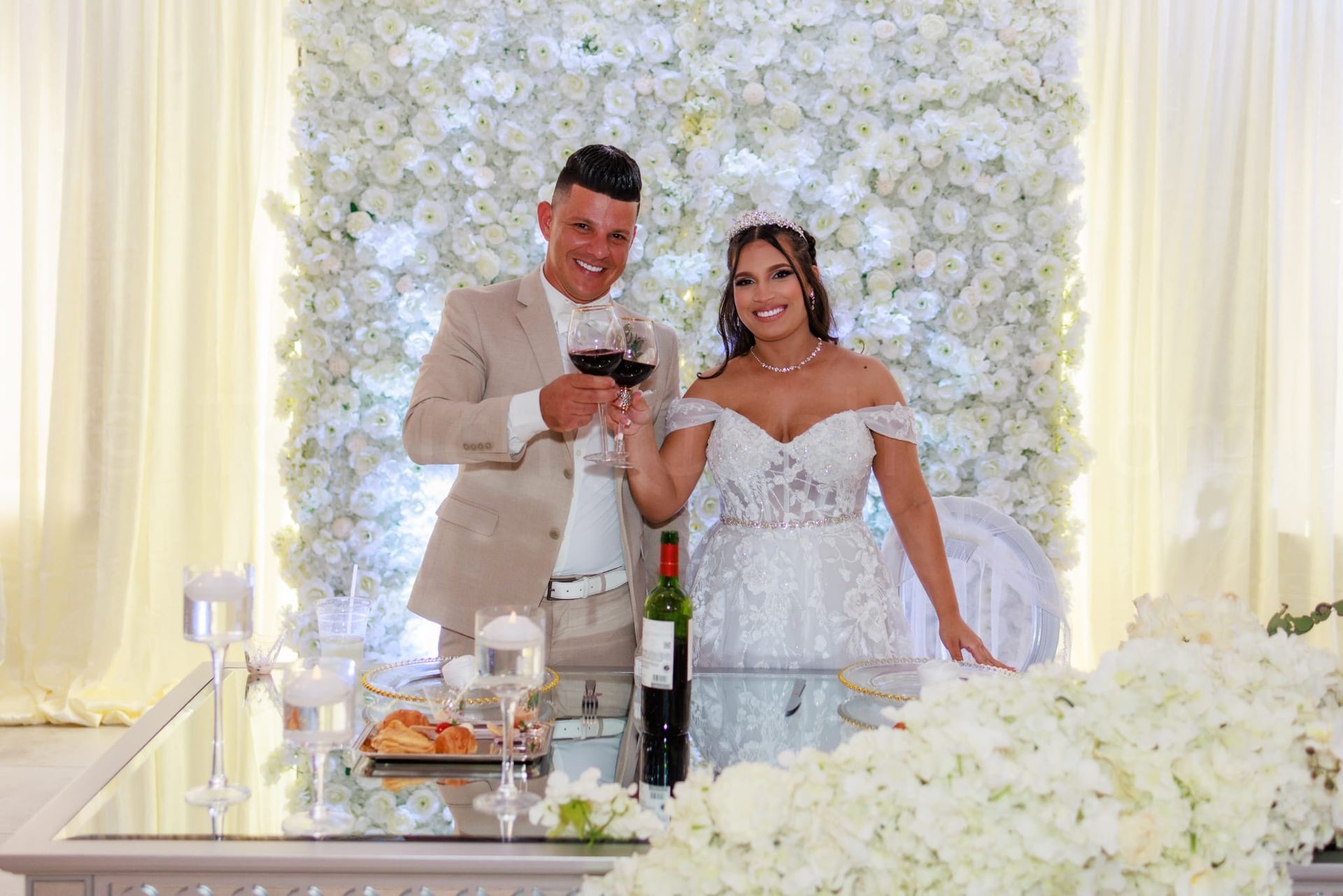 Couple toasting with wine at a wedding reception, set against a wall of white flowers.