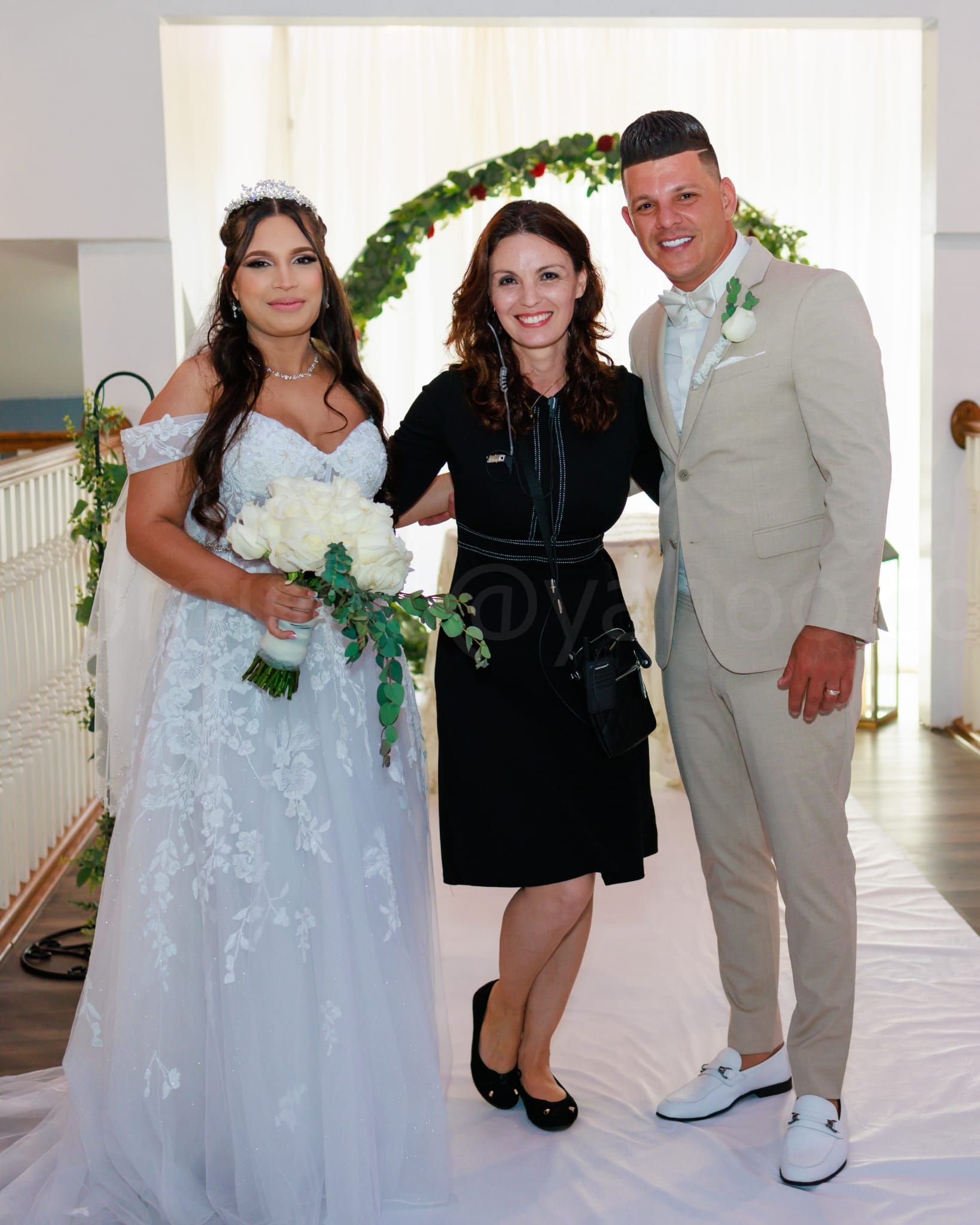 Bride and groom pose with wedding planner under floral archway. Bride wears white gown; groom in light suit.