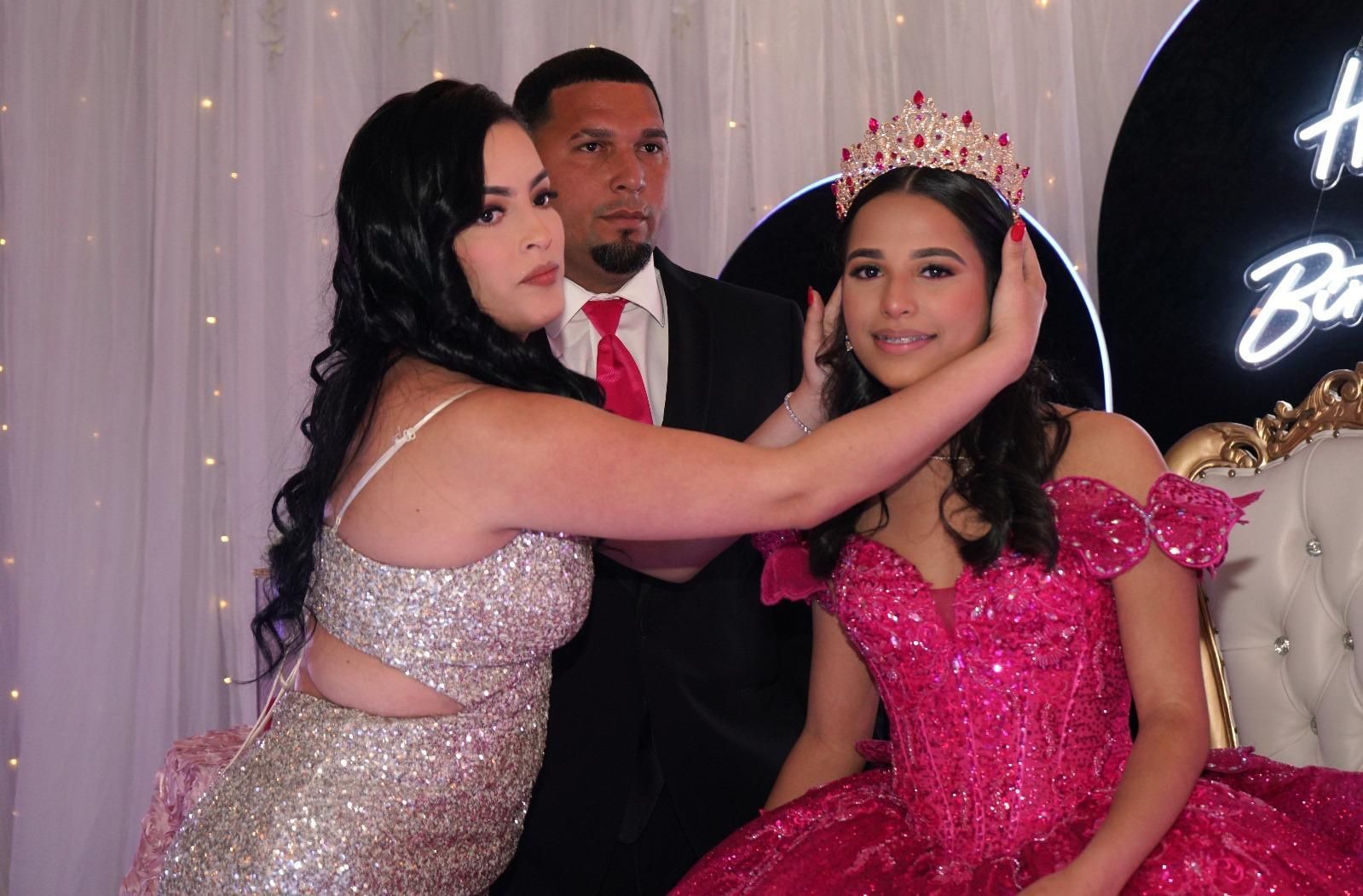 Woman places a crown on another woman's head, while a man in a suit looks on. Festive backdrop.
