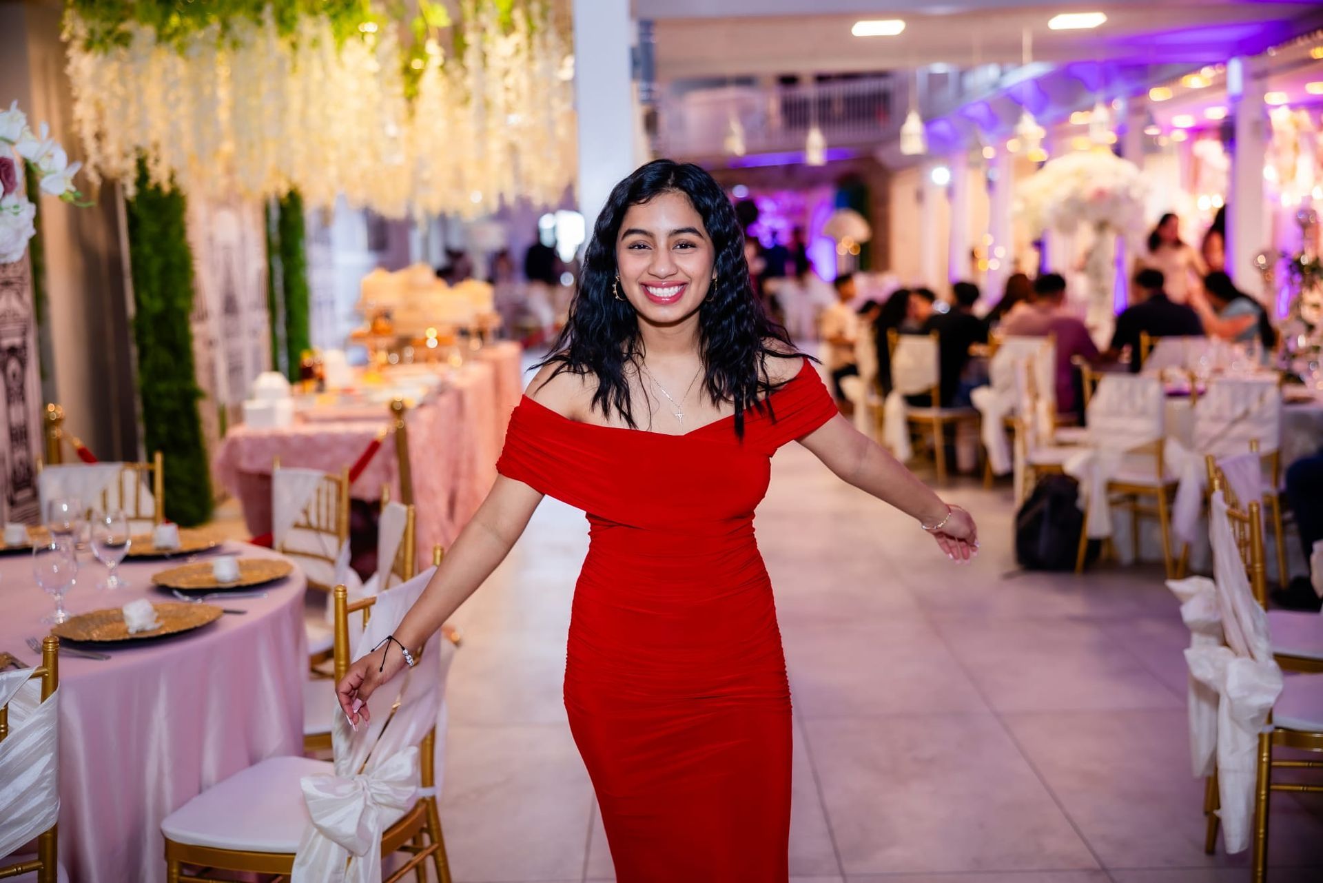 Woman in a red off-shoulder dress smiles at a formal event. Tables set with flowers, white chairs.