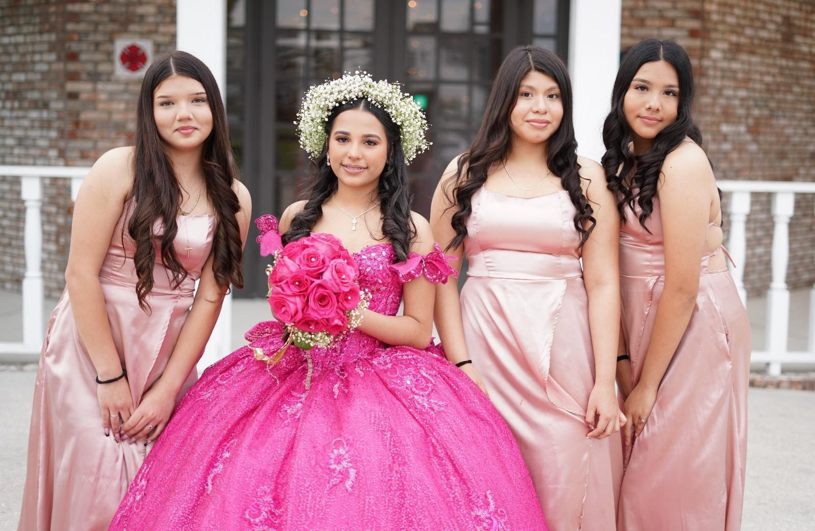 Group of young women pose outdoors; the center person wears a bright pink gown and flower crown, holding pink roses; others in pink dresses.