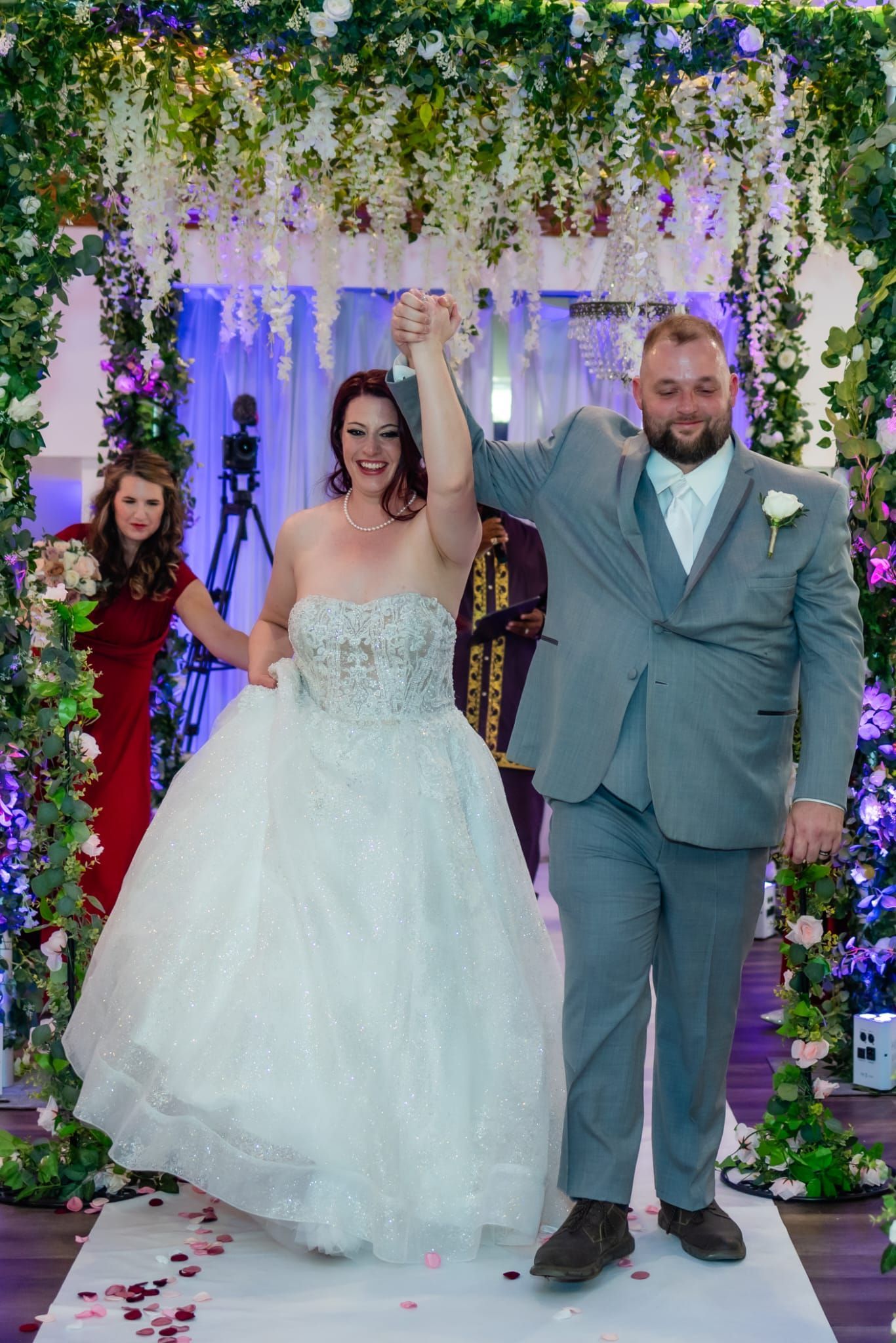 Newly married couple exiting a wedding ceremony under an arch of flowers, woman in a white gown and man in a grey suit.