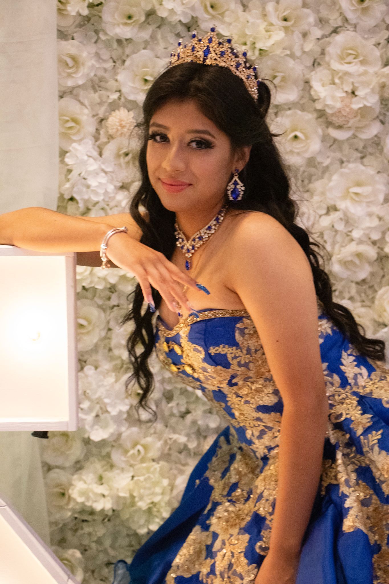 Woman in blue and gold dress, crown, jewelry; smiling, leaning on a light, in front of a white floral wall.